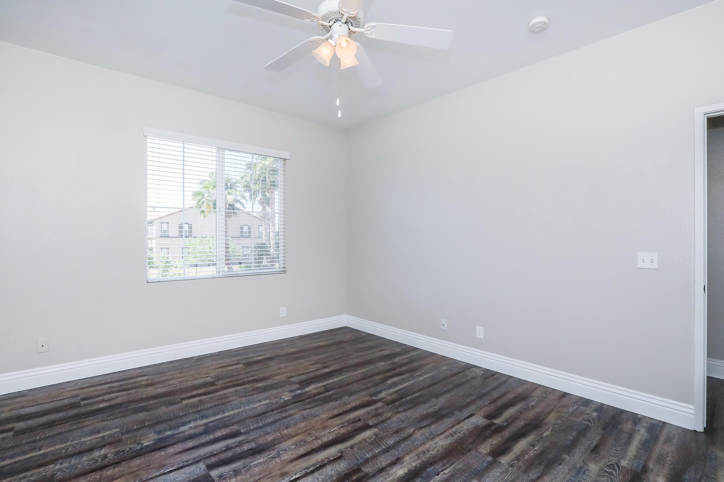 A spacious, empty room with light-colored walls and dark wooden flooring. A ceiling fan with light hangs in the center, and a window with blinds allows natural light to enter, showcasing palm trees outside. The room features a door on the right leading to another space.