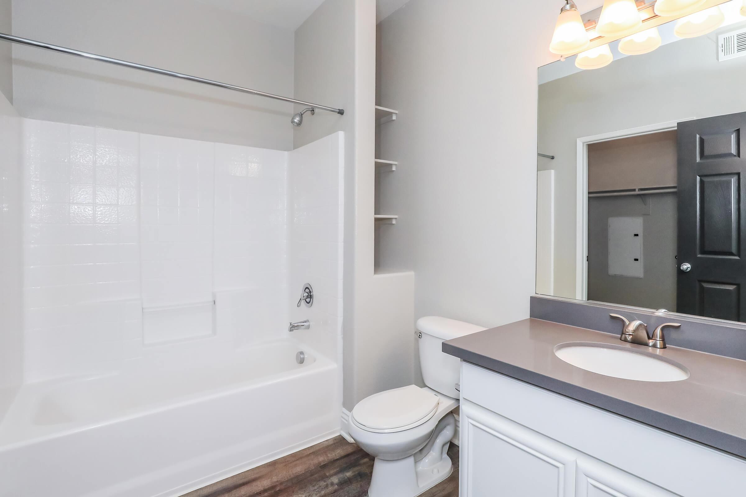 A modern bathroom featuring a white bathtub with a shower, a toilet, and a vanity with a sink. The walls are a light color, and there are shelves next to the tub. A large mirror above the sink reflects the space, and soft lighting hangs from the ceiling. The flooring is a wood-like laminate.