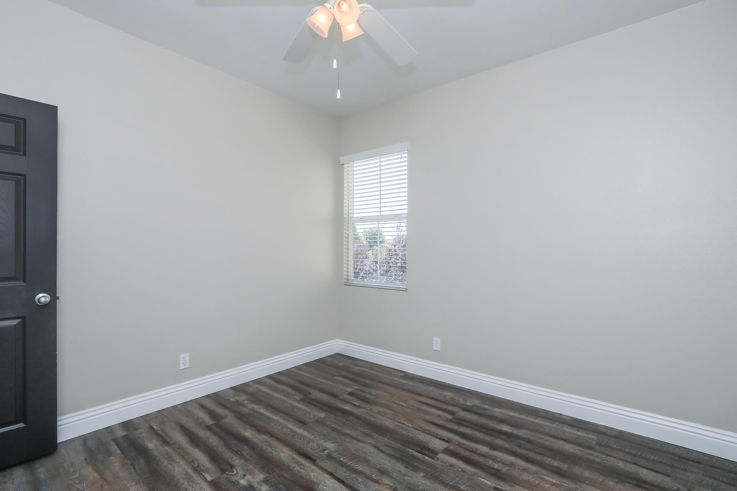 Empty room featuring light gray walls, a ceiling fan, a window with white blinds, and dark wood-like flooring. A black door is visible on the left. The space is well-lit, with natural light coming through the window.