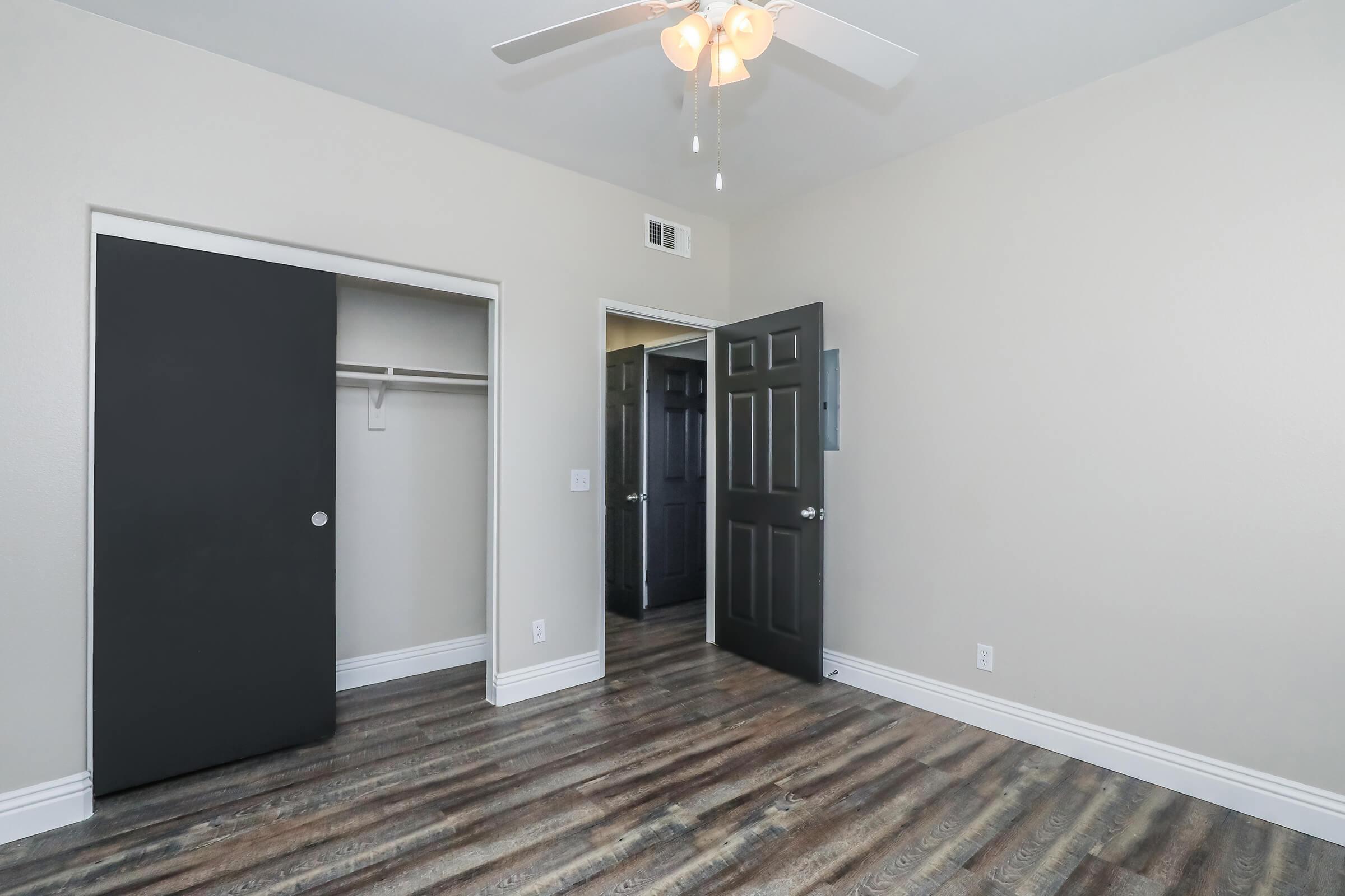 An interior view of a small bedroom featuring light-colored walls, a ceiling fan, and a wood-like floor. There are two doors: one leading to a closet and another open door suggesting an adjacent room. The overall aesthetic is modern and clean, with ample natural light.