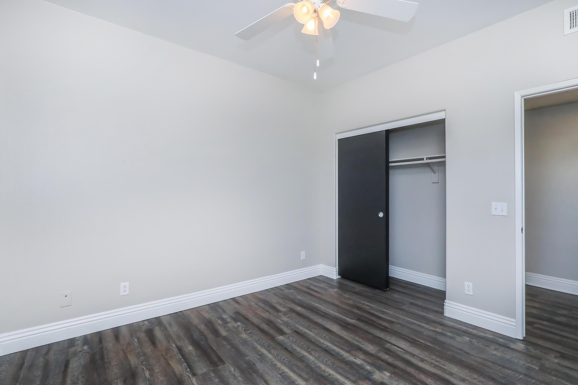 A spacious, empty bedroom featuring light-colored walls and a ceiling fan. There is a black sliding closet door on one side, and a door leading to another room on the opposite side. The floor is covered with dark laminate wood flooring, creating a modern and clean aesthetic.