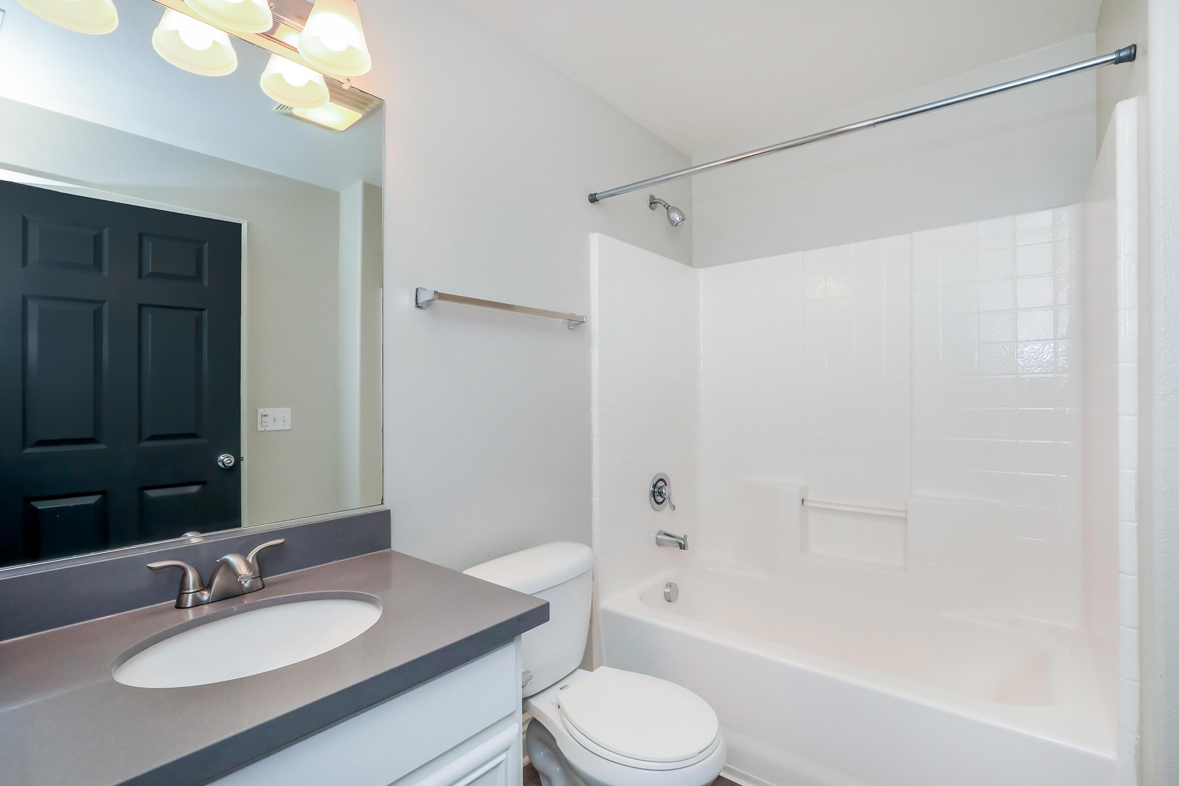A clean and modern bathroom featuring a white bathtub and shower combination, a wall-mounted mirror above a sink with a gray countertop, and a dark door in the background. The lighting is soft, coming from overhead fixtures. The overall color scheme is neutral and bright.