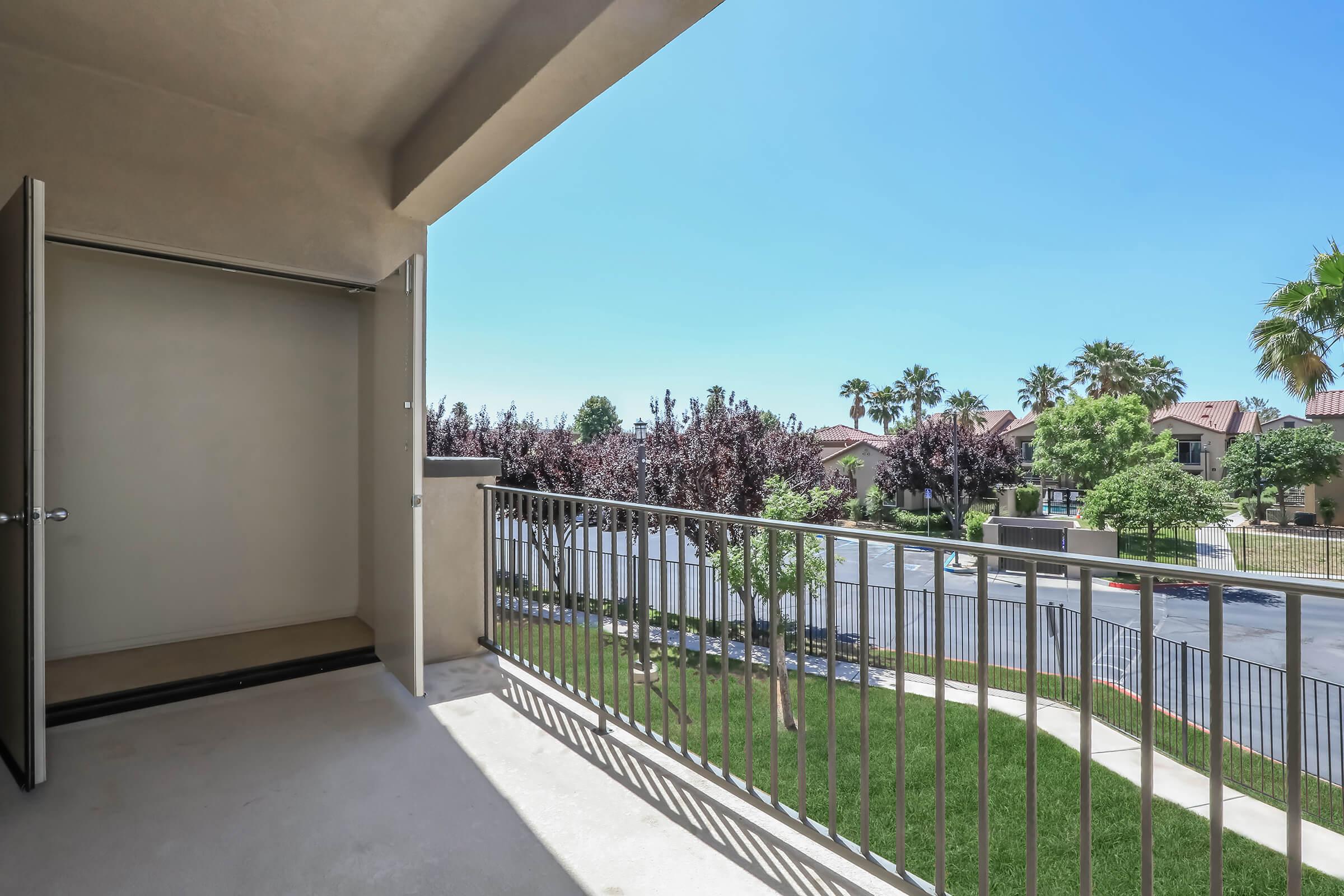 View from a balcony overlooking a landscaped area with trees and a parking lot. The sky is clear and blue, and nearby buildings are visible in the background. The balcony features a railing and an open door leading to an interior space.