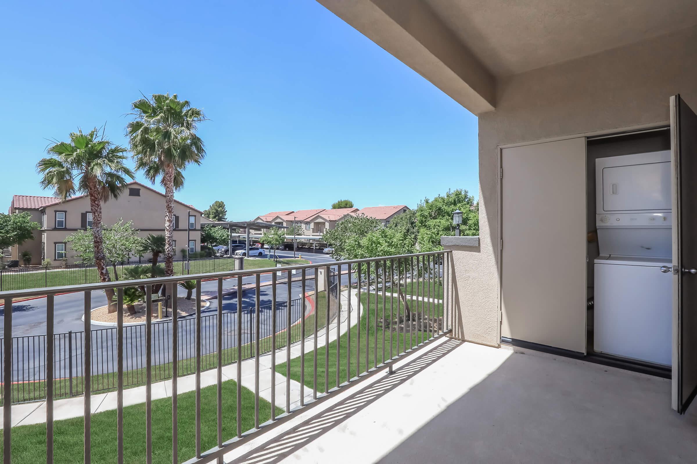 View from a balcony overlooking a landscaped area, featuring palm trees and neighboring buildings. A doorway leads to a laundry area with a stacked washer and dryer. The sky is clear and blue, creating a bright and inviting atmosphere.