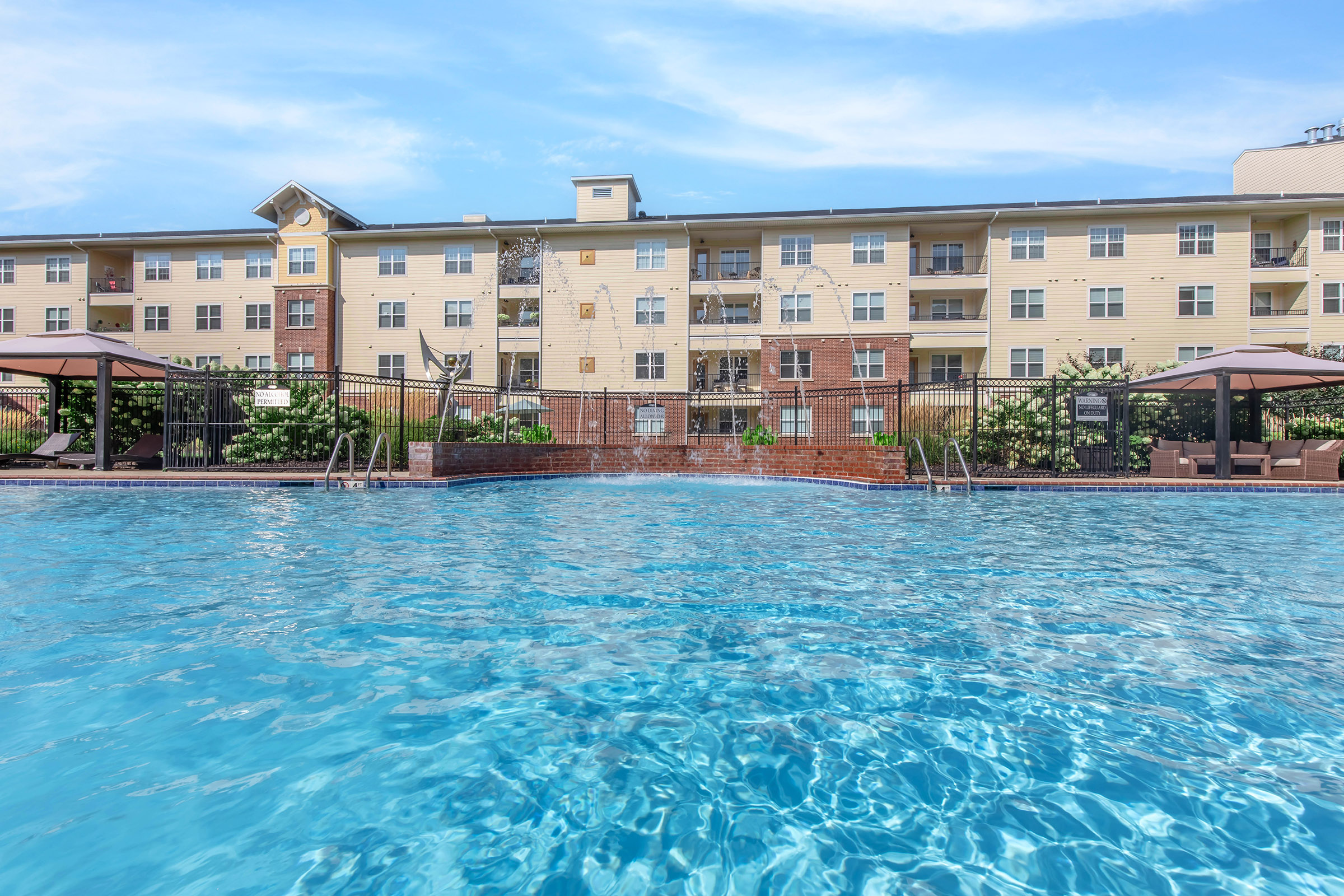 A clear swimming pool in the foreground with a vibrant blue surface, reflecting the sky. In the background, a multi-story apartment building with balconies is visible, surrounded by landscaped greenery and lounge areas with shade structures. The scene conveys a relaxed, inviting atmosphere.
