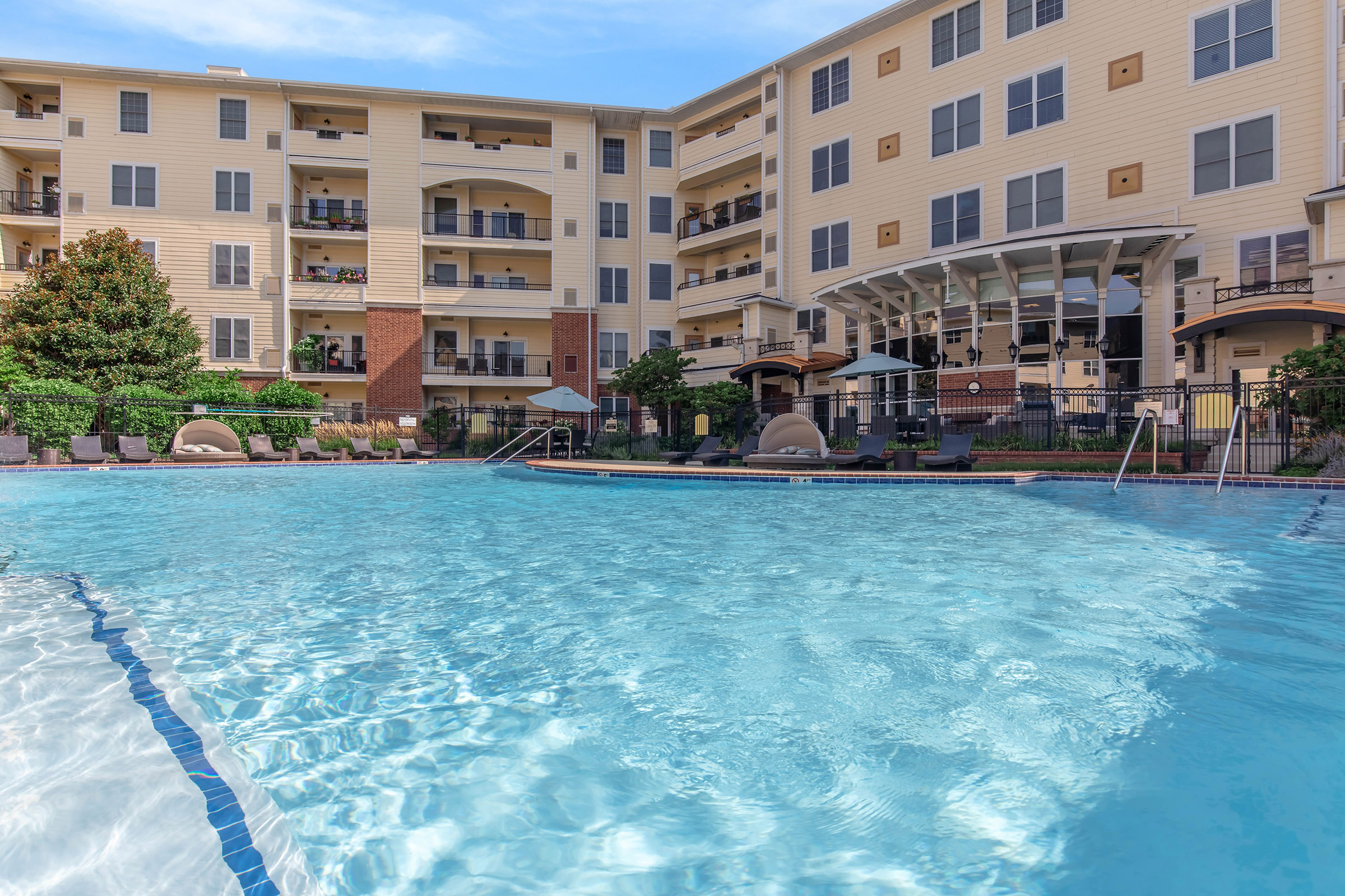 A clear blue swimming pool surrounded by lounge chairs and umbrellas, with a residential building featuring balconies in the background. Lush greenery enhances the pool area, creating a relaxing and inviting atmosphere.