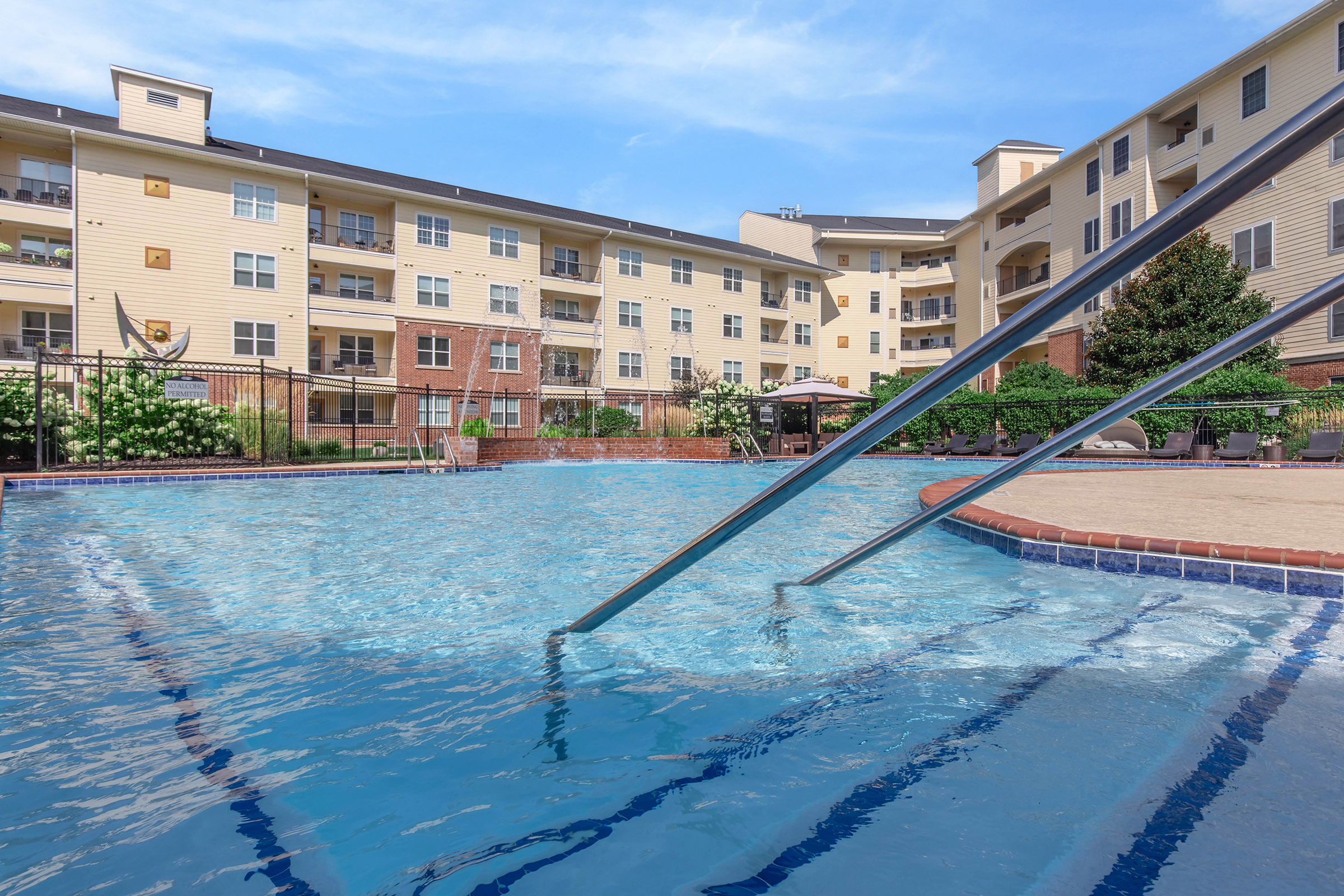 Swimming pool with clear blue water and metal handrails, surrounded by landscaped greenery and a multi-story apartment building in the background. The sky is partly cloudy, creating a serene atmosphere. Sun loungers and umbrellas are visible near the pool area.