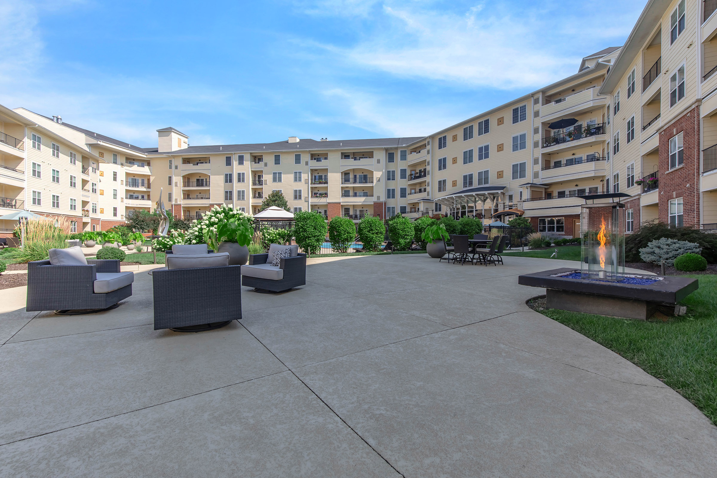 A well-designed outdoor area featuring modern seating arrangements with two gray chairs and a fire pit. Lush green landscaping surrounds the space, along with a pathway and elegant buildings in the background under a clear blue sky. Perfect for relaxation and social gatherings.