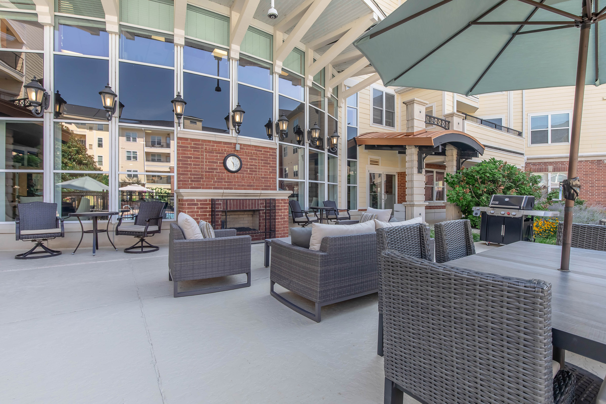 A cozy outdoor seating area featuring stylish wicker furniture, a clock on the wall, and a barbecue grill. Large windows allow plenty of natural light, and the space is decorated with potted plants, creating a welcoming environment for relaxation or social gatherings.
