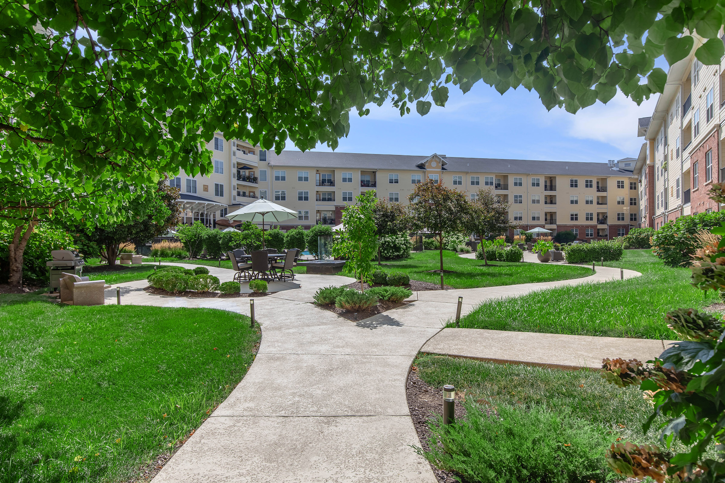 A landscaped garden with a winding pathway surrounded by lush green grass and neatly trimmed bushes. In the background, an apartment building with balconies is visible. Patio furniture under an umbrella is set up for outdoor relaxation, creating a serene and inviting atmosphere.