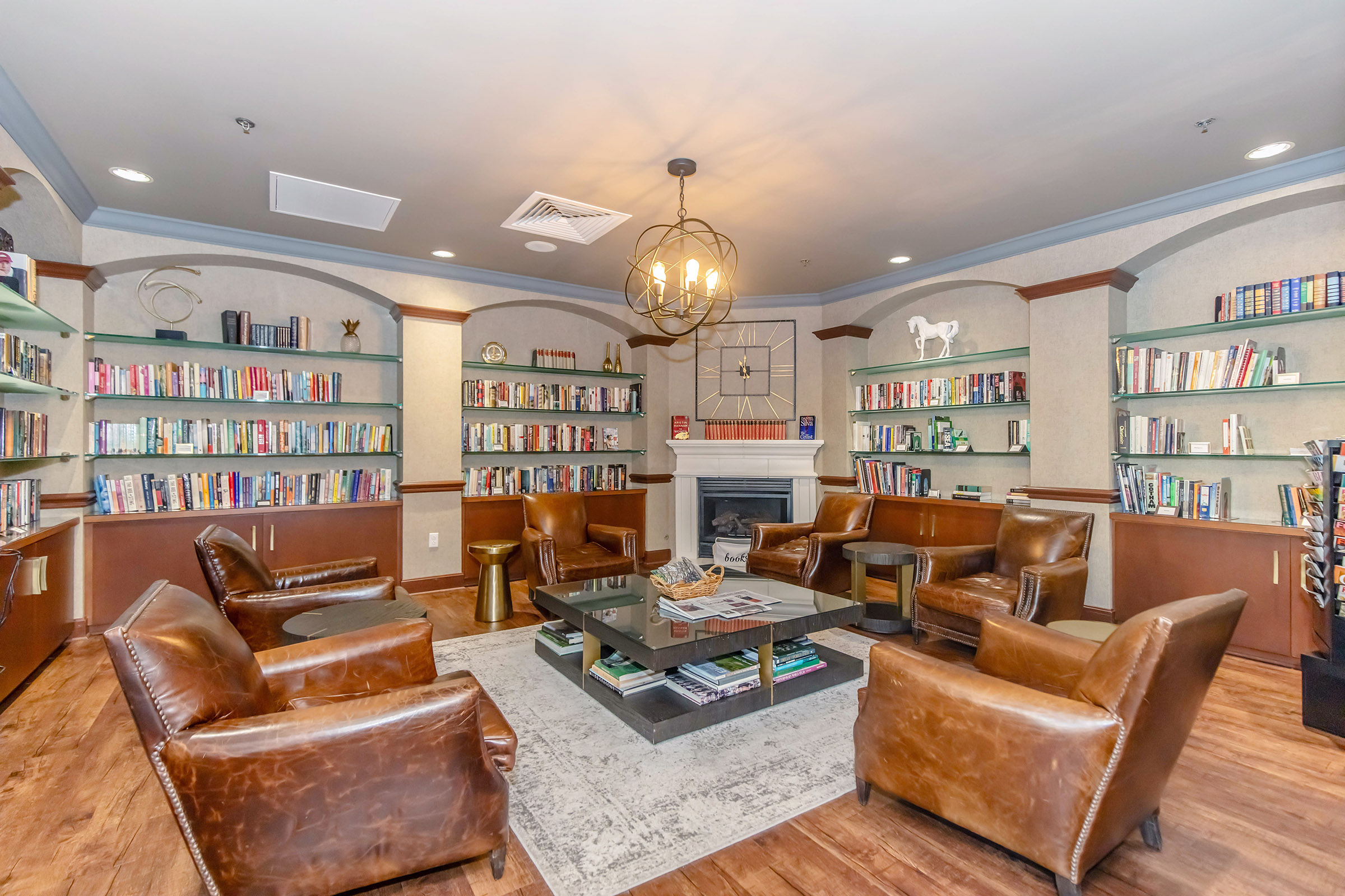 Cozy reading room featuring leather armchairs arranged around a central table, surrounded by bookshelves filled with books. A fireplace adds warmth to the space, complemented by decorative items on the shelves and a stylish light fixture overhead. Ideal for relaxation and reading.