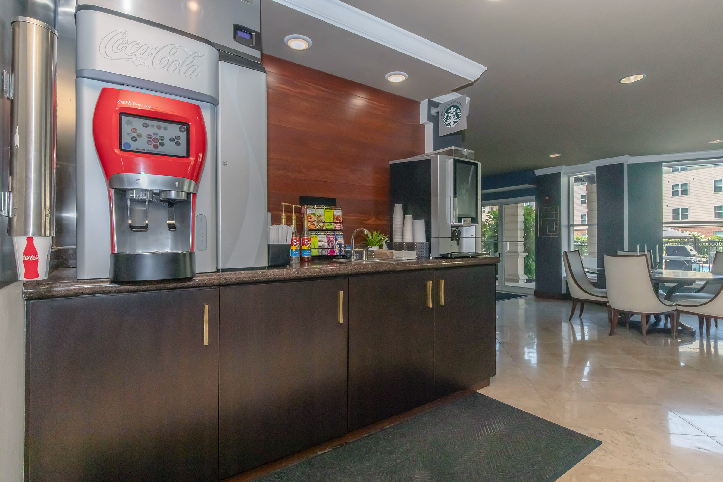 A modern refreshment area featuring a Coca-Cola soda fountain machine, various beverage options, and sleek cabinetry. The space has a contemporary design with a view of dining tables and outdoor scenery in the background, creating a welcoming atmosphere for guests.