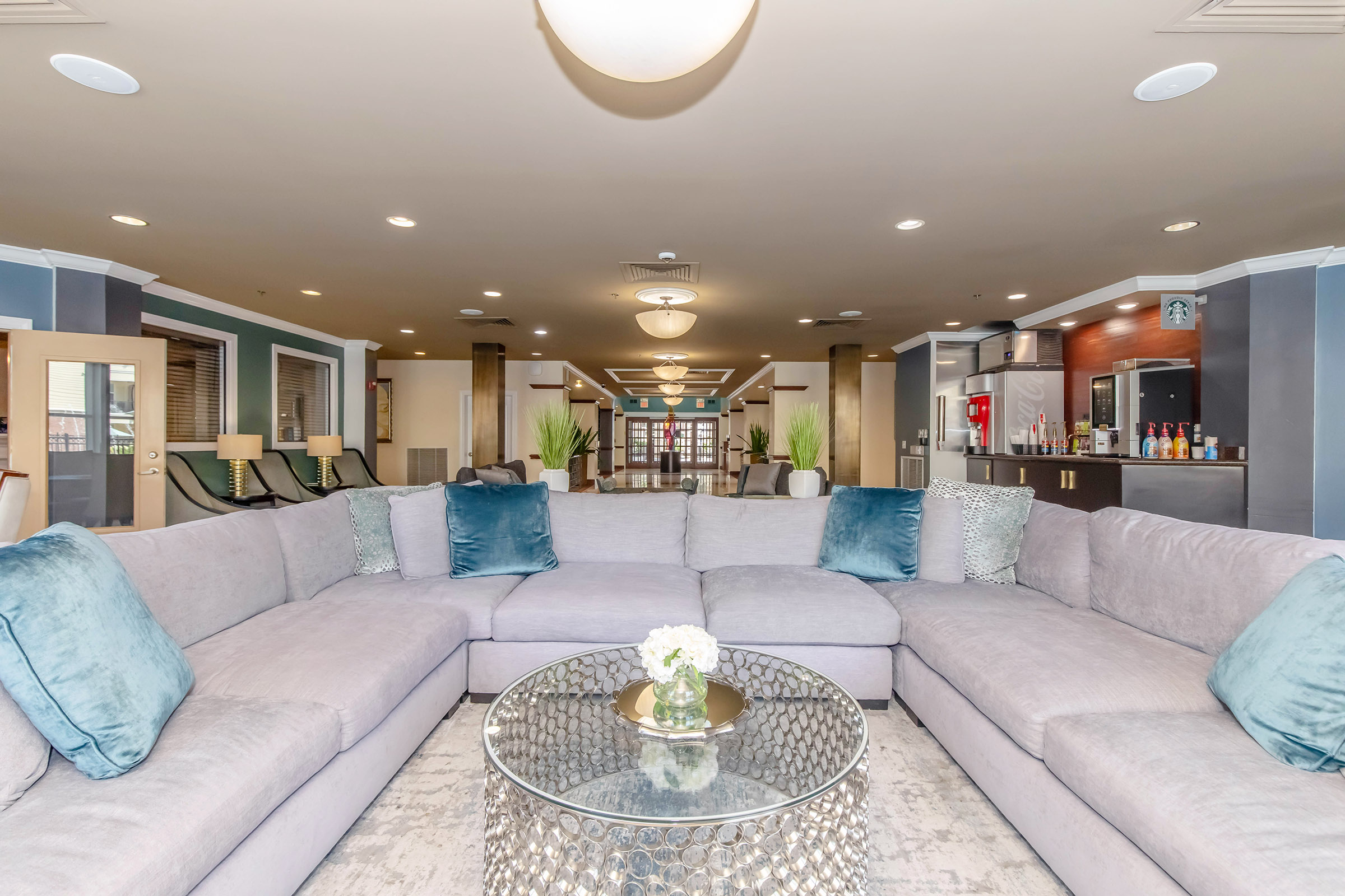 A spacious lounge area featuring a large, curved grey sectional sofa adorned with blue cushions. In the center, there's a glass coffee table with a small vase of flowers. The background shows a well-lit reception area with modern decor, plants, and a bar-like counter.