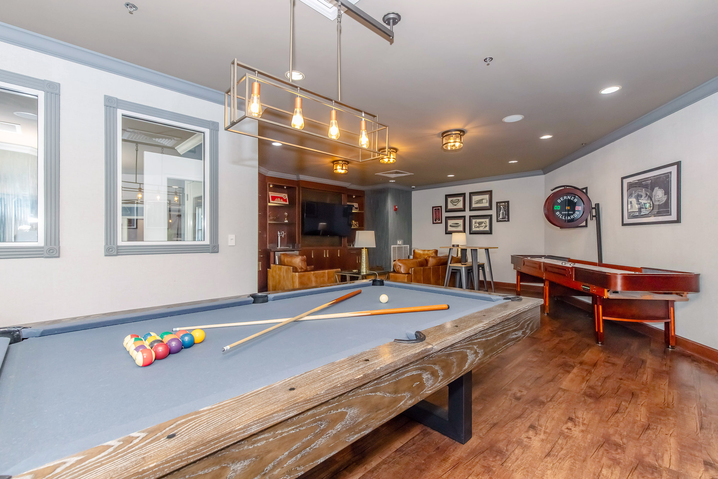 A modern game room featuring a billiards table with cues and balls, a wooden floor, and stylish armchairs. The room is well-lit with hanging pendant lights and decorated with framed artwork. In the background, there is a TV and a bar table with stools, creating a relaxed atmosphere for entertainment.
