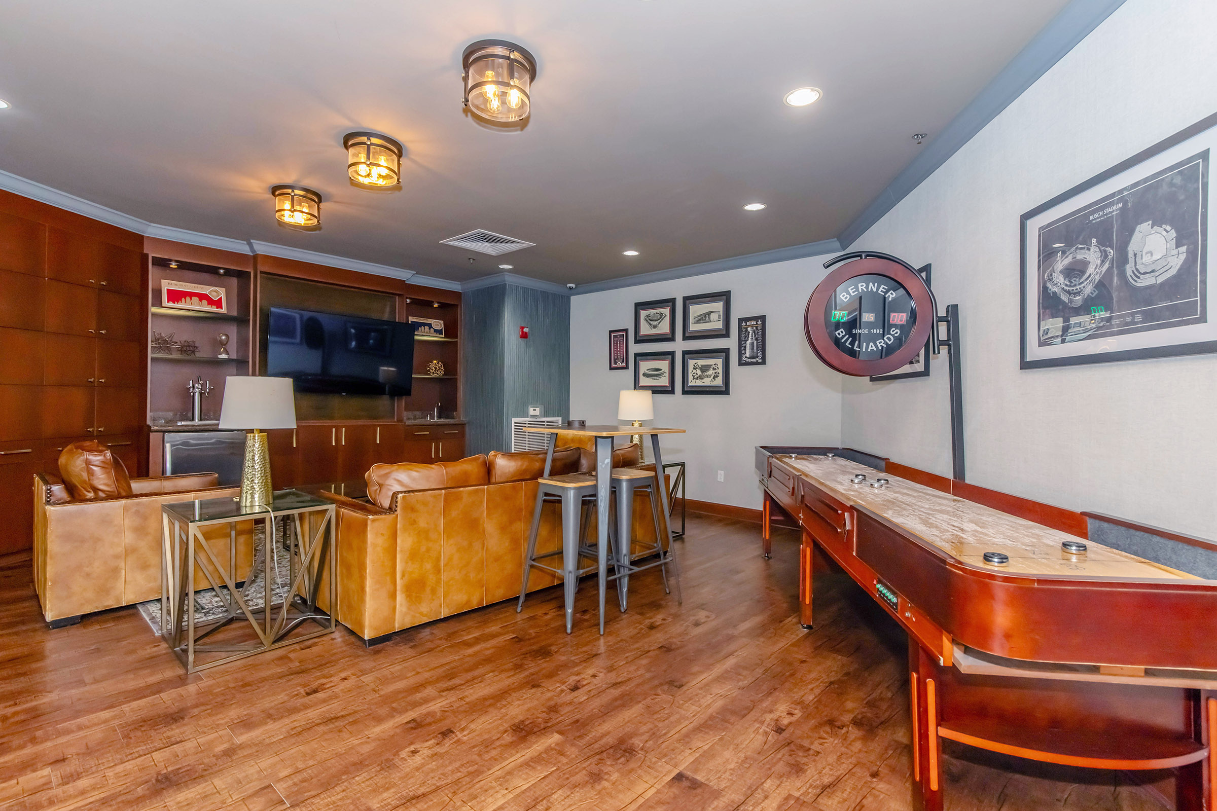 A cozy game room featuring a shuffleboard table, leather seating, and stylish lighting. The walls are adorned with framed art and photographs, contributing to a warm and inviting atmosphere. There is a television and a bookshelf in the background, completing the recreational space.
