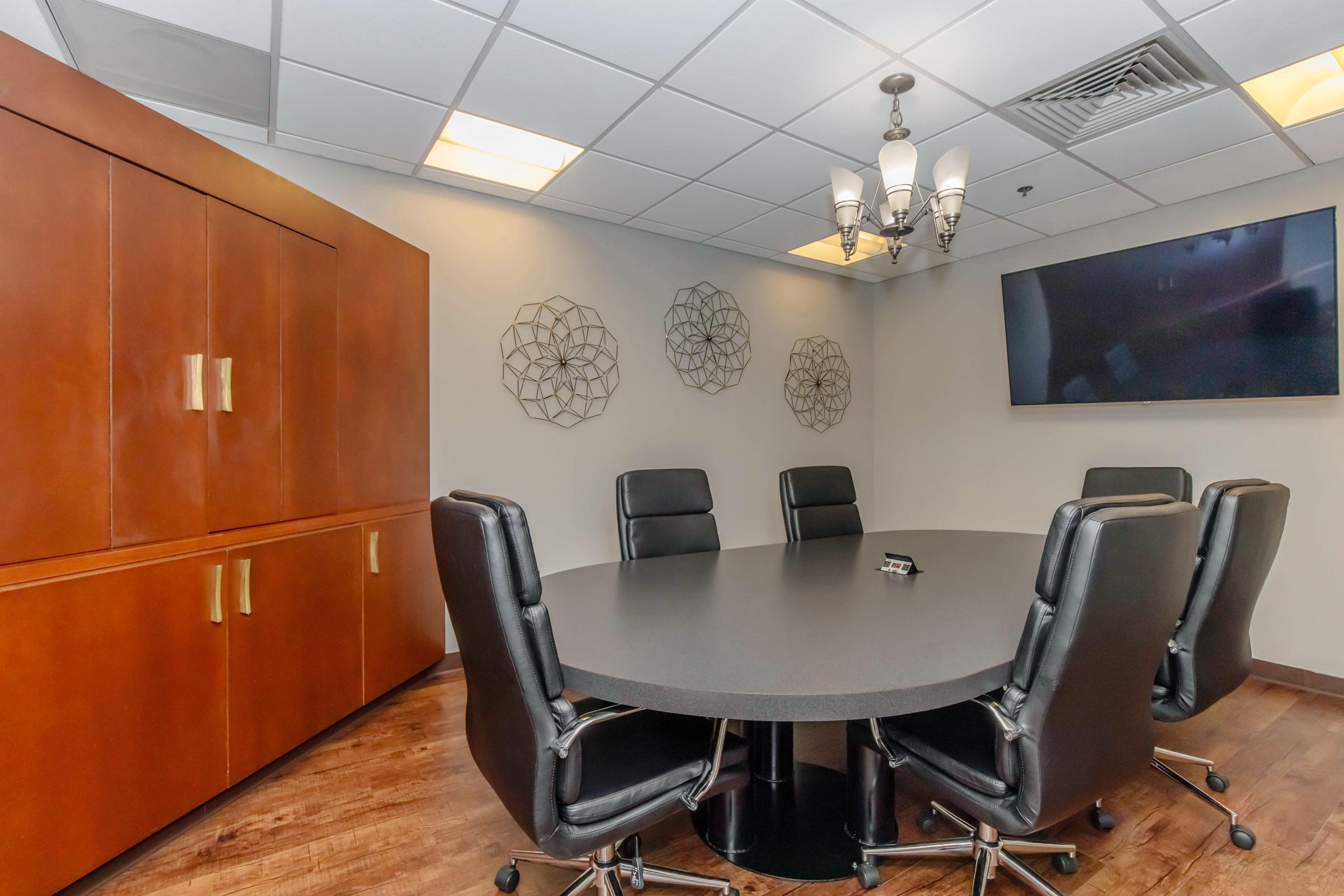 A modern conference room featuring a round black table surrounded by black leather chairs. The walls have decorative metal wall art, and there is a large flat-screen TV mounted on one wall. The room has warm lighting and a wooden cabinet for storage.