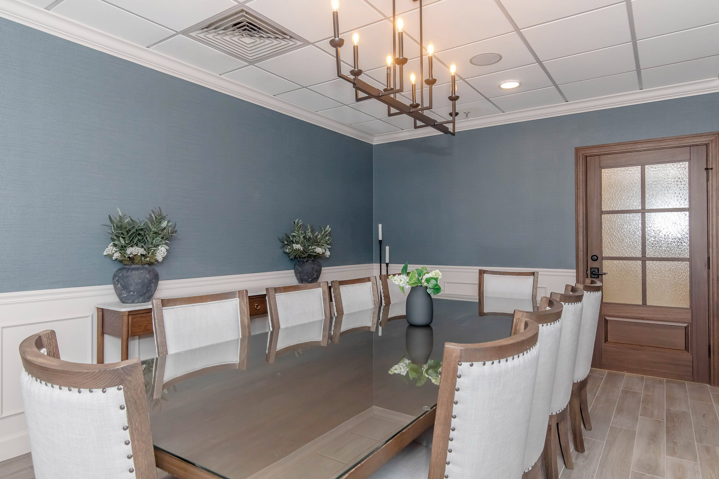 A modern conference room featuring a large glass table surrounded by white-upholstered chairs. The walls are painted in a soft blue shade, and two small potted plants are placed on the table. A chandelier hangs from the ceiling, and a wooden door with a glass panel is visible in the background.