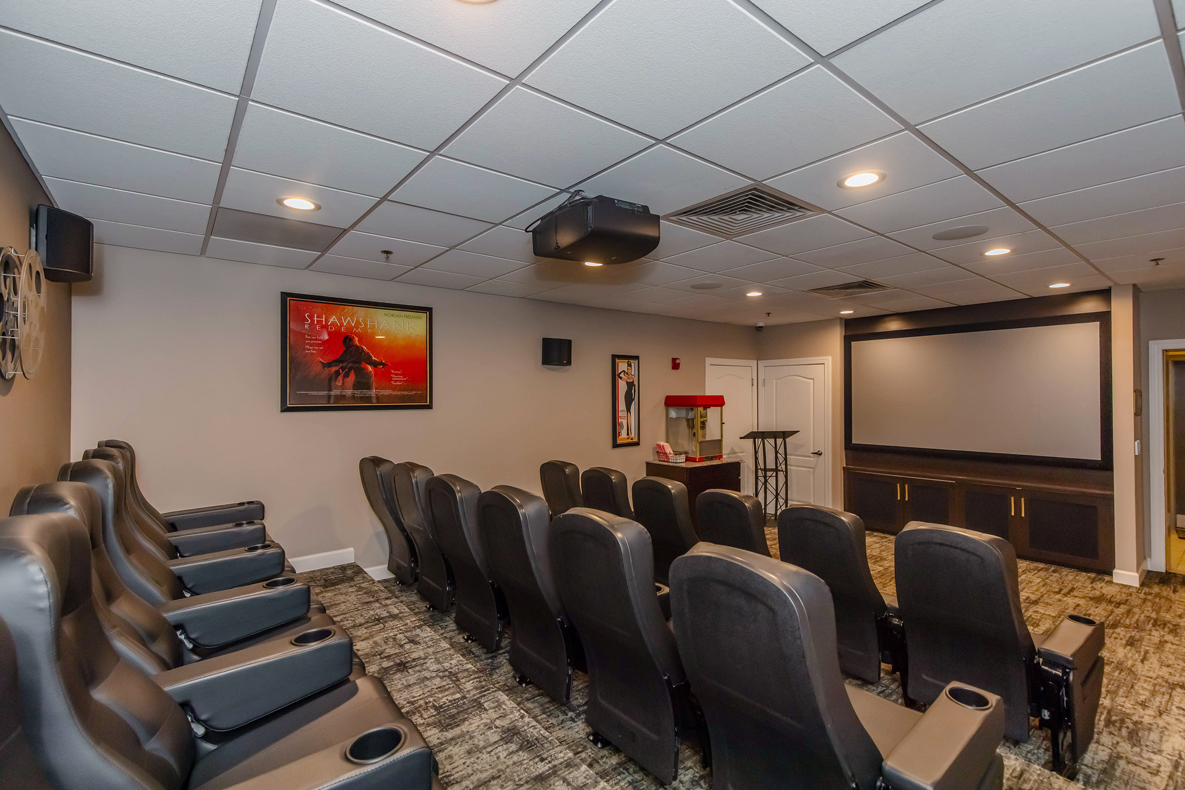 A home theater room featuring comfortable black seating arranged in rows, with a large screen and projector mounted on the ceiling. The walls are painted in a neutral tone, and there is a poster hanging for "Shawshank Redemption." A vintage popcorn machine adds a classic touch to the space.