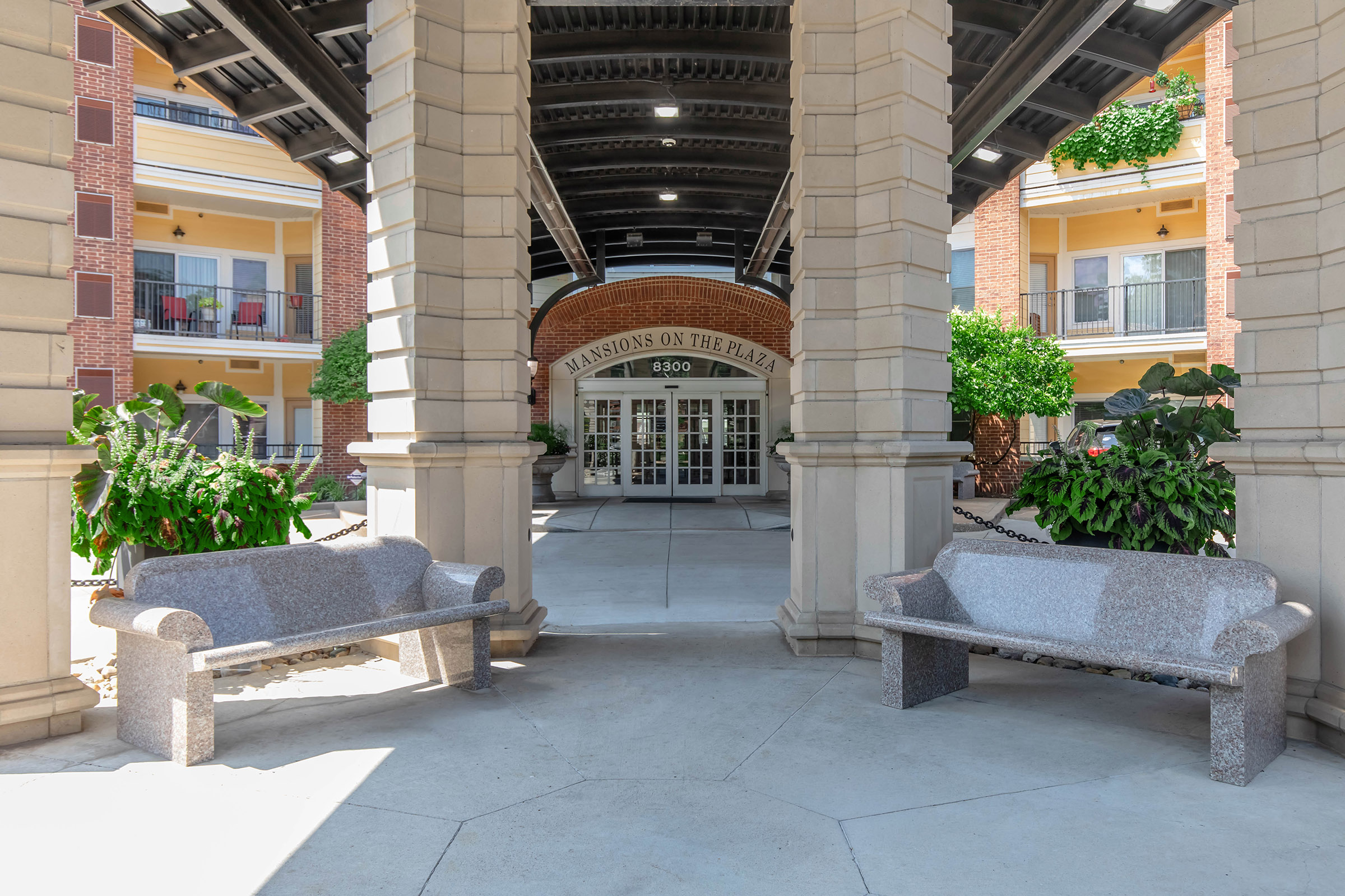 A view of the entrance to "Mansions on the Plaza," featuring a spacious atrium with stone archways. Two gray stone benches are positioned on either side, surrounded by lush greenery and potted plants. The double glass doors lead into a welcoming lobby area, enhancing the inviting atmosphere.