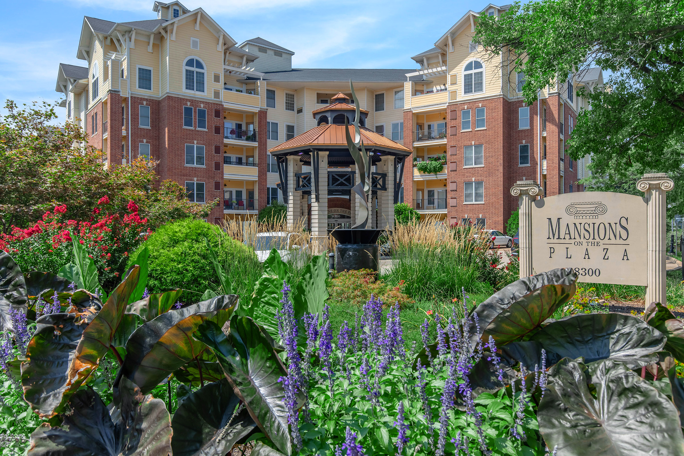 A well-maintained apartment complex with a gazebo and lush landscaping featuring colorful flowers and plants. The sign reads "Mansions in the Plaza," indicating the name of the property. The building is multi-storied with brick and beige siding, surrounded by greenery.