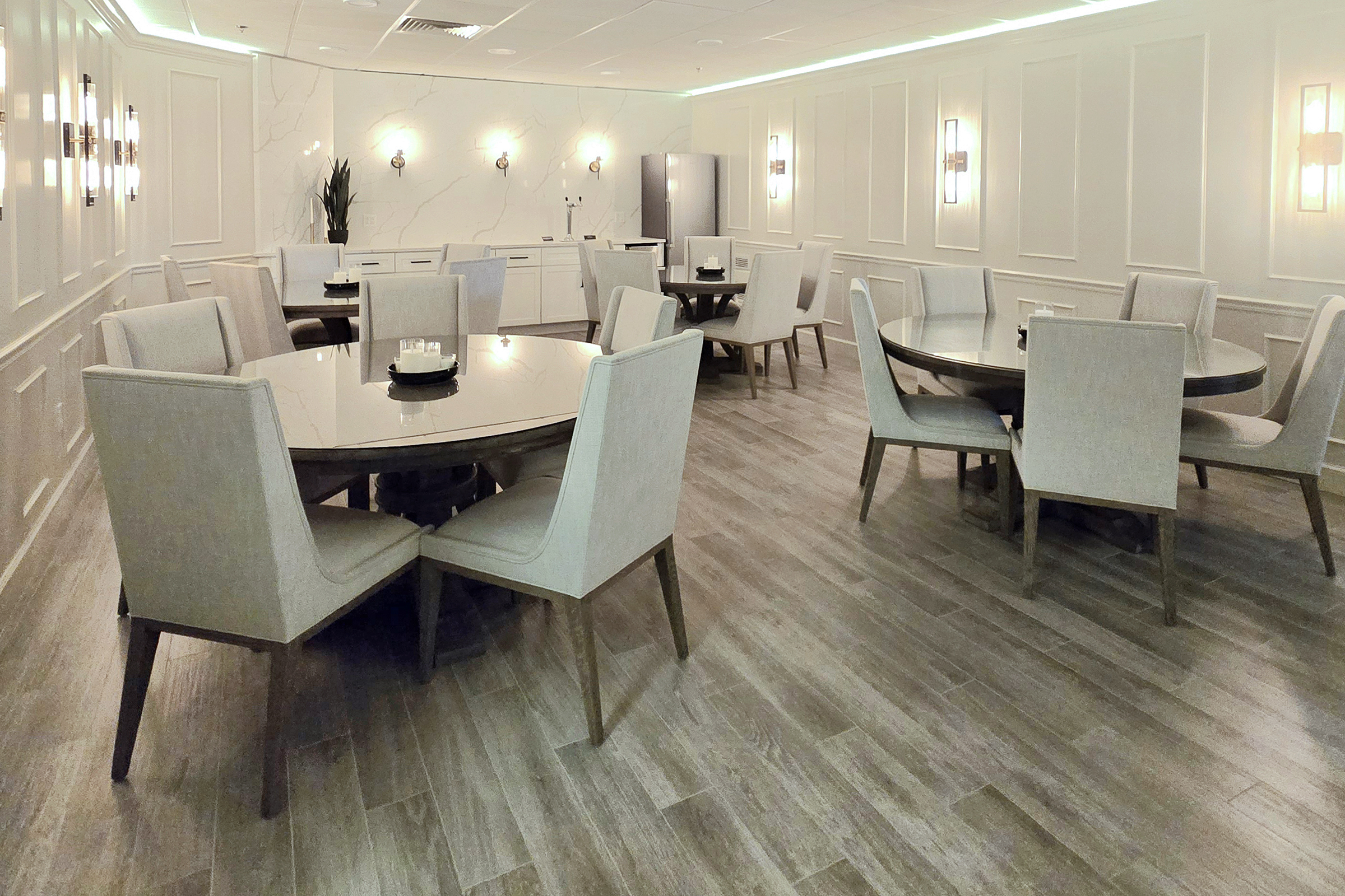 A modern dining area featuring multiple circular tables surrounded by light-colored upholstered chairs. The floor is made of wood-like tiles, and the walls are adorned with subtle paneling and lighting fixtures, creating a bright and inviting atmosphere. A plant is visible in the background.