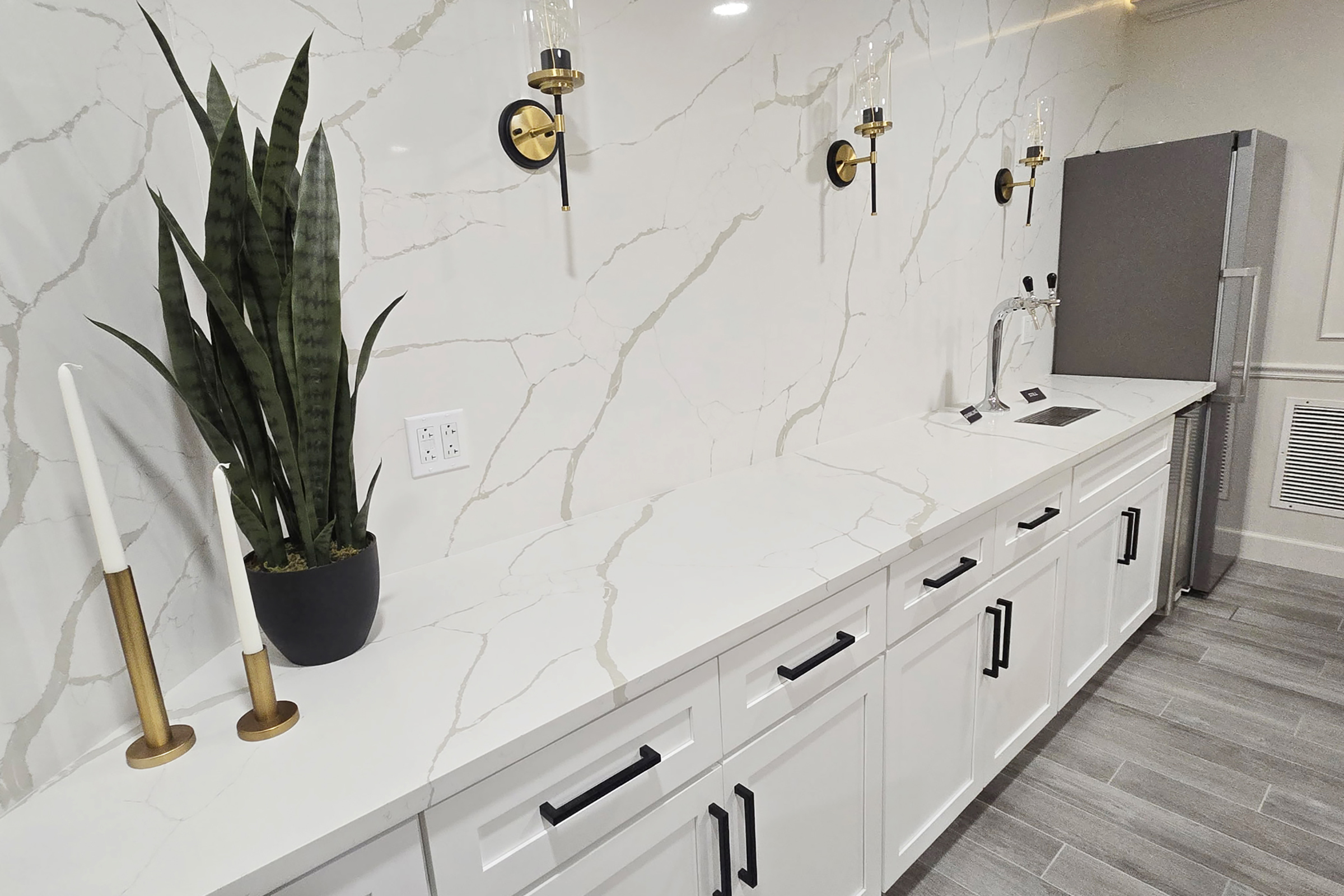 A modern kitchen counter featuring a sleek white marble surface with gold accents. Two wall-mounted sconces provide lighting, while a tall green plant in a black pot adds a touch of nature. The white cabinets below have black handles, and a stainless steel refrigerator is visible in the background.