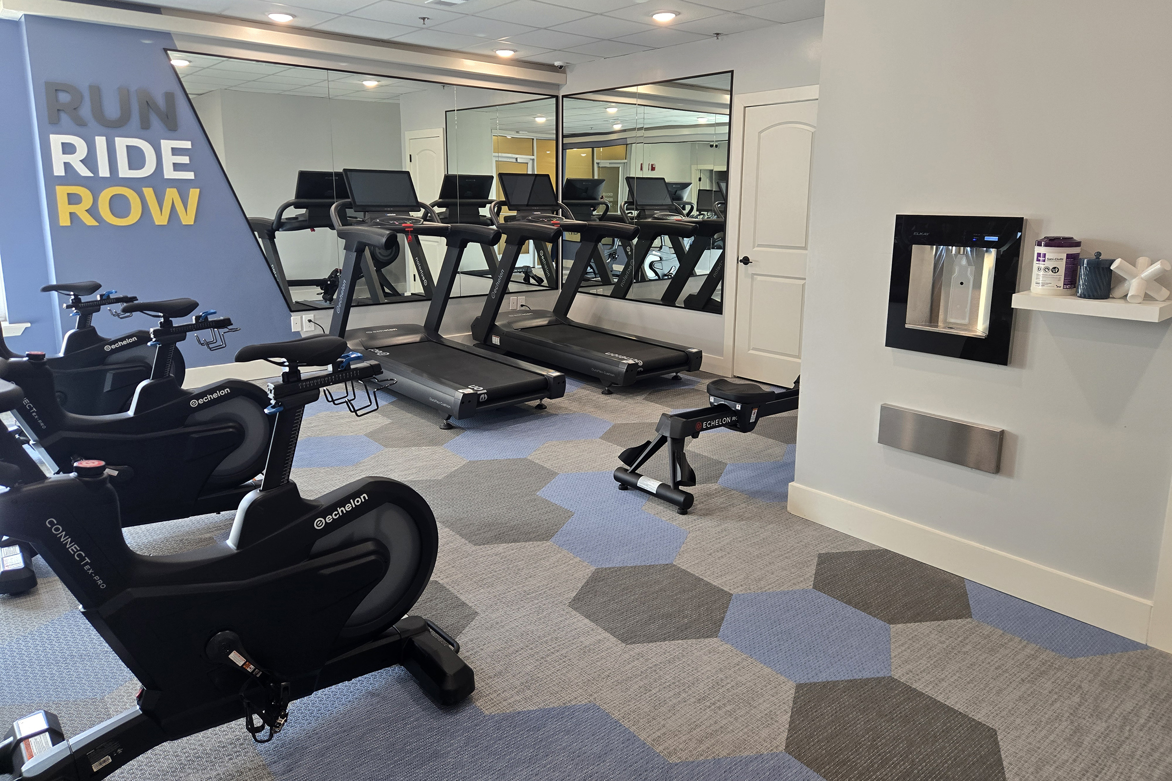 A modern fitness room featuring several treadmills and stationary bikes. Gray and blue hexagonal carpet tiles cover the floor. A mirrored wall reflects the equipment, and there is a water fountain on the wall nearby. The room is bright and designed for exercise, with motivational phrases displayed.