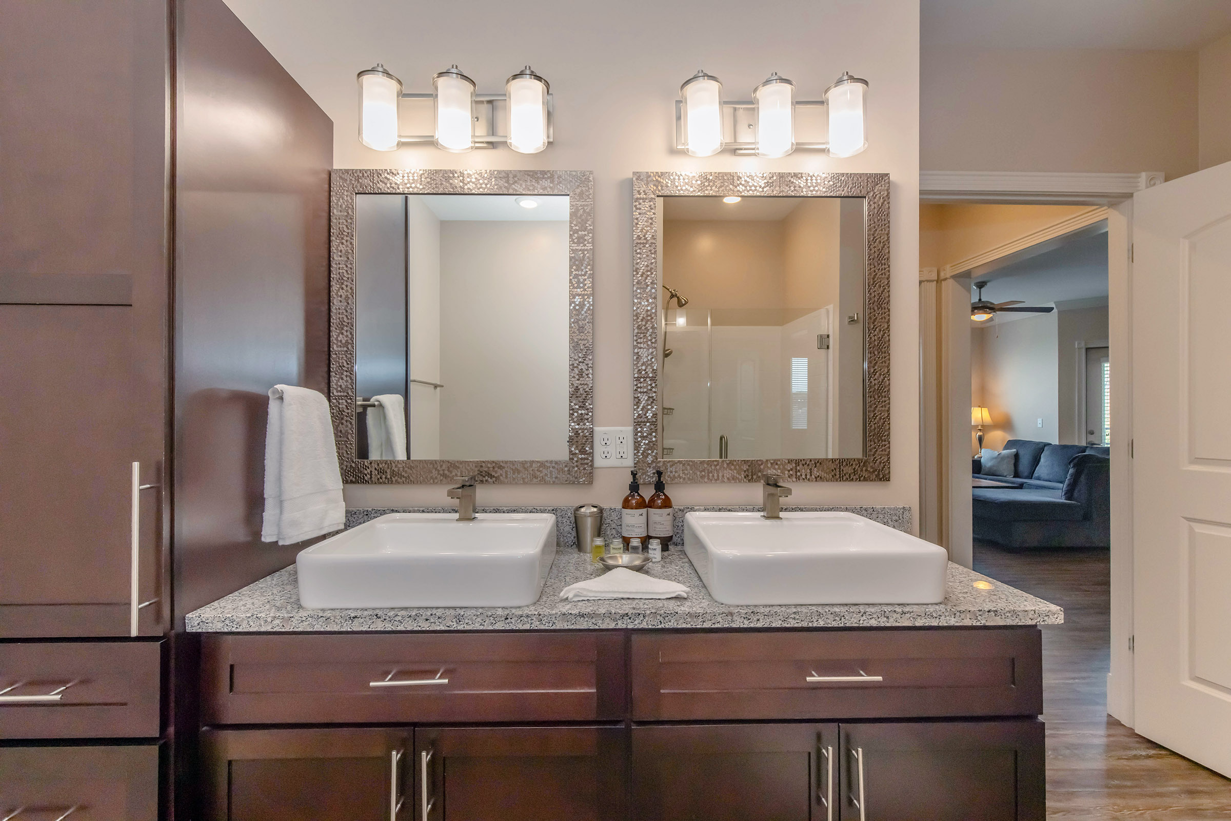 A modern bathroom featuring double sinks with rectangular white basins, a stylish granite countertop, and decorative mirrors above each sink. Bright overhead lights illuminate the space, while a towel hangs on the side. In the background, a glimpse of a cozy living room is visible through an open doorway.