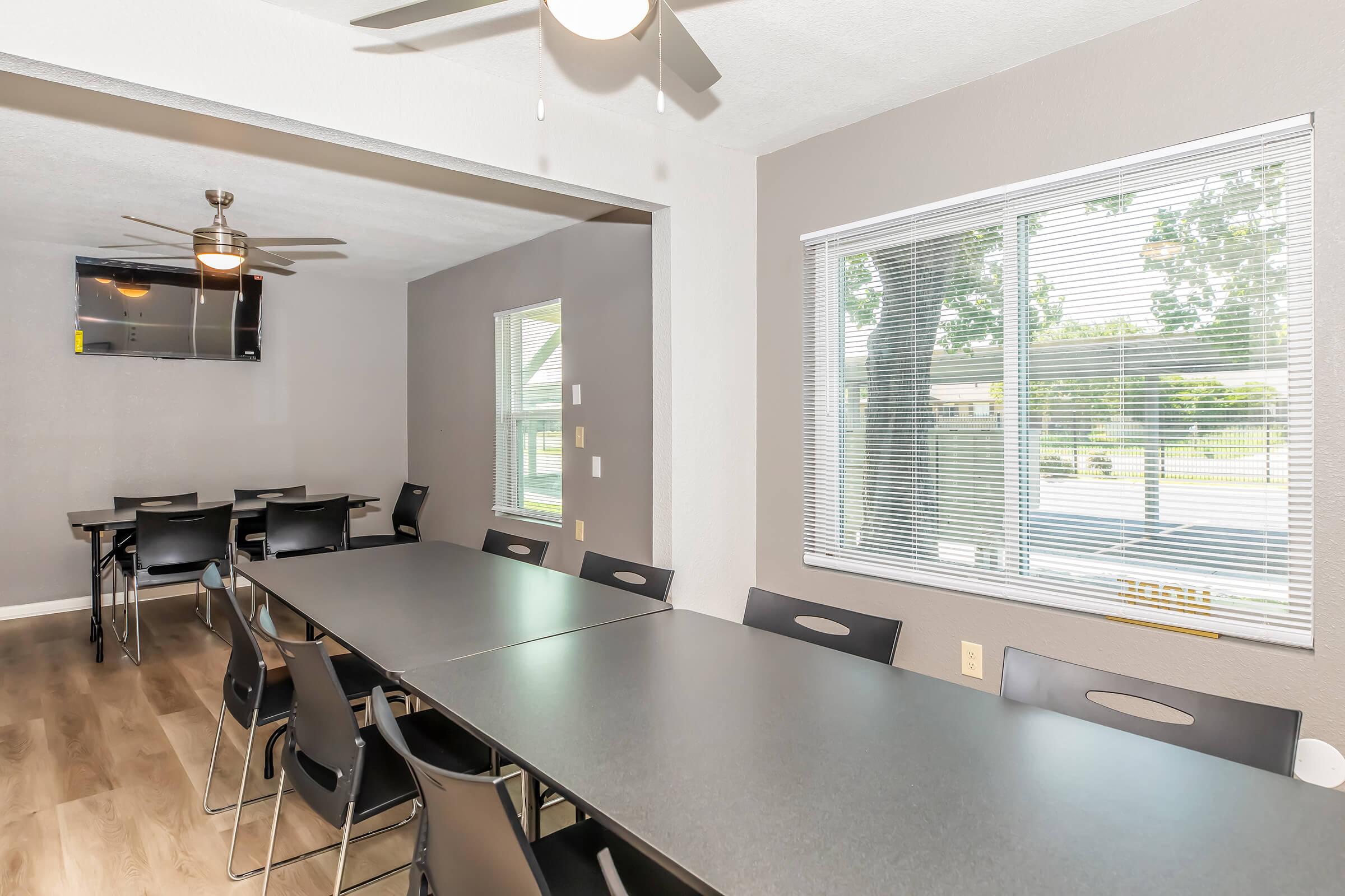 A spacious meeting room featuring a long table with black chairs, windows providing natural light, and a mounted television. The walls are painted in a neutral color, and ceiling fans enhance the comfort of the space. Ideal for gatherings or presentations.