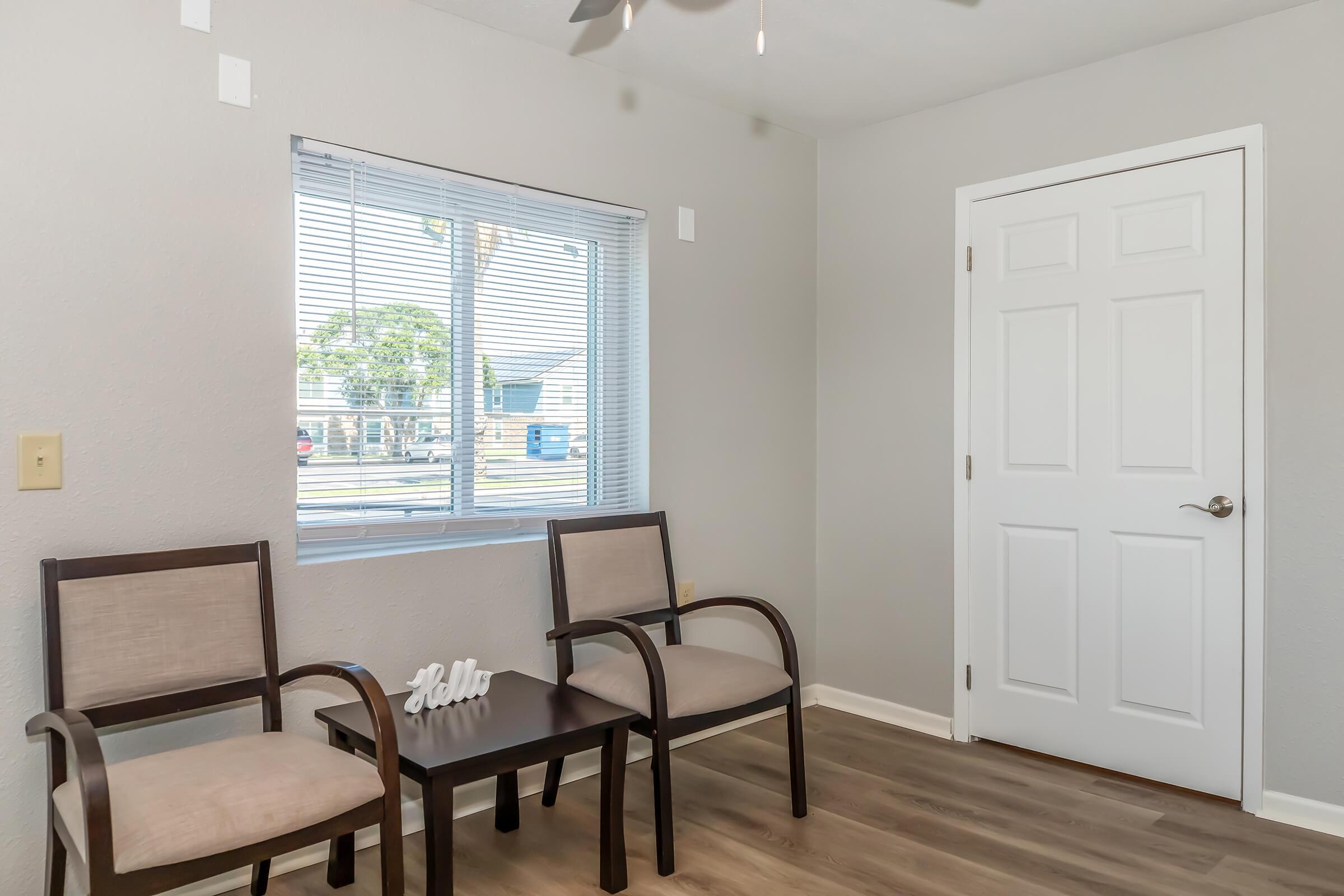 A bright, modern room featuring two wooden chairs on either side of a small table. A window with blinds allows natural light to enter, while a closed white door is visible on the right. The walls are painted a soft gray, and the floor is a light wood finish, creating a welcoming atmosphere.