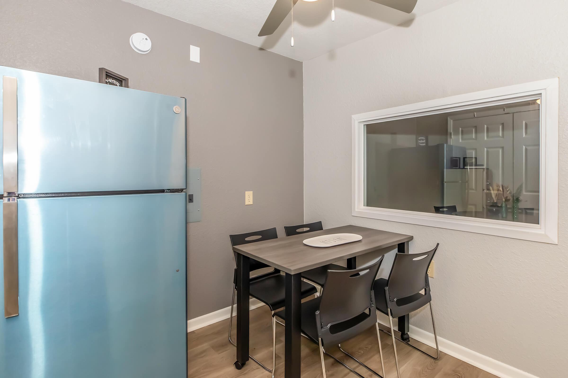 Modern kitchen setting featuring a stainless steel refrigerator and a small dining table with four black chairs. The wall is painted in a light gray tone, enhancing the contemporary feel. A window provides a view to another room, adding natural light to the space.