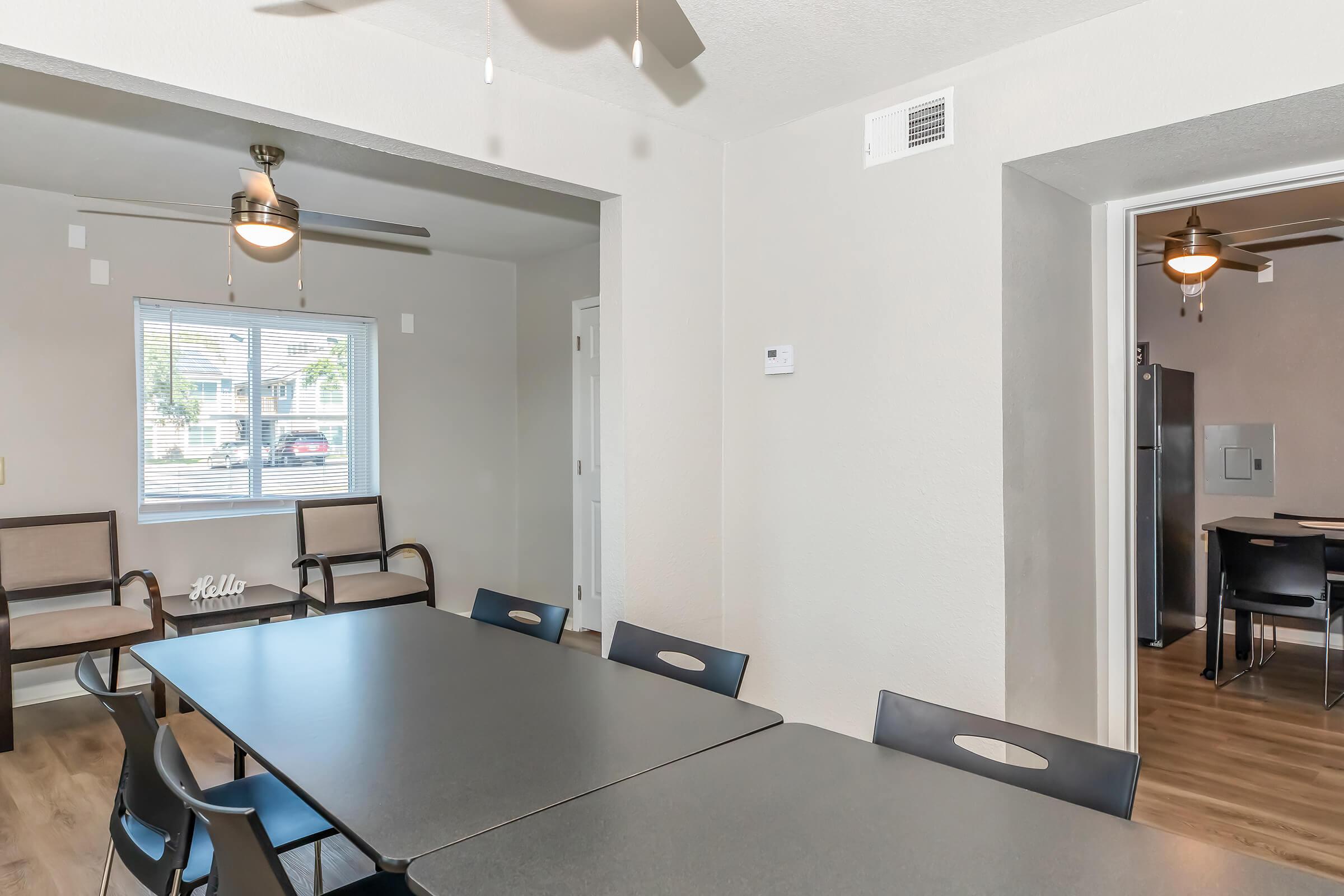 A spacious room with a large black table surrounded by black chairs. There are two ceiling fans overhead, and a window allowing natural light. In the background, a doorway leads to another area with a refrigerator and additional seating. The walls are light-colored, creating a bright, airy atmosphere.