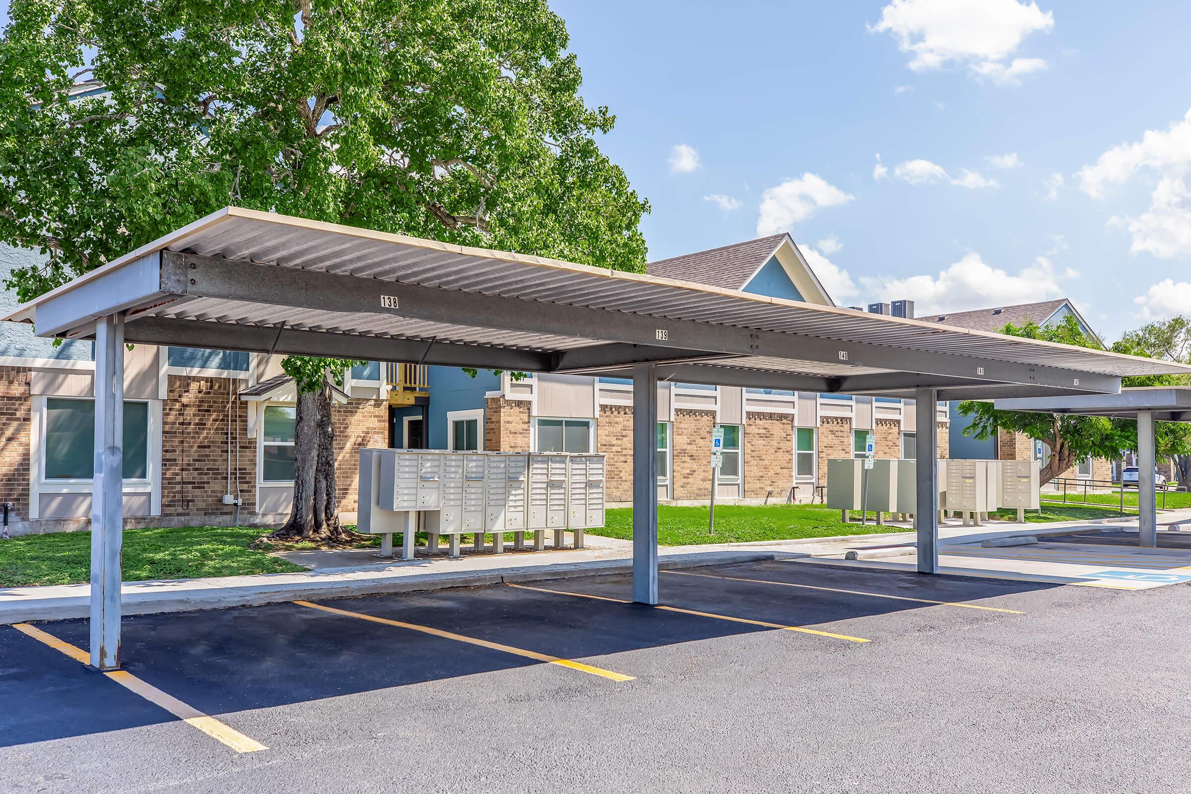 A covered parking area with several empty spaces beneath a large awning, beside a residential building. In the background, there are mailboxes aligned next to the parking area, with green grass and trees surrounding the scene under a clear blue sky.