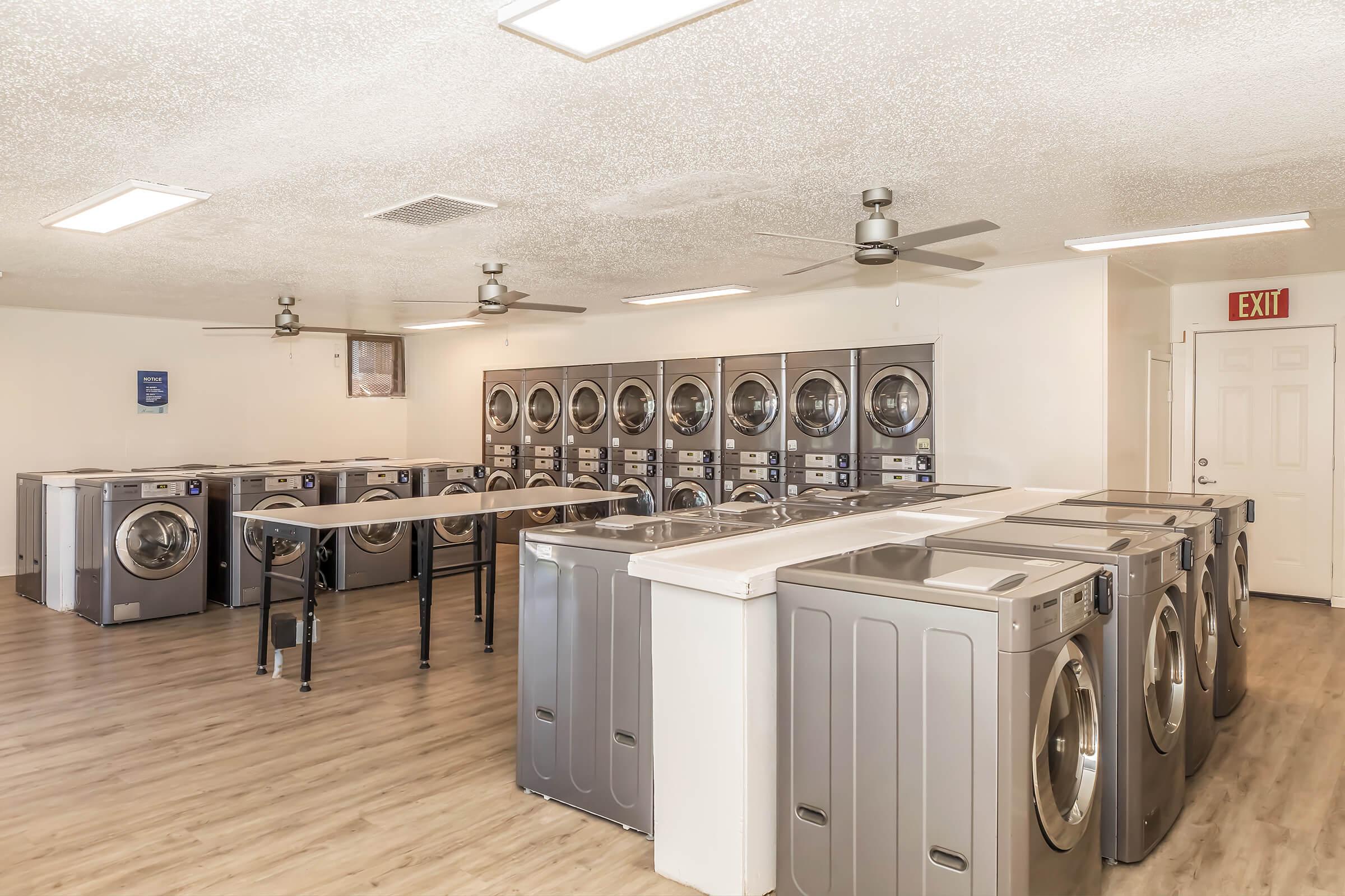 A modern laundry room featuring several rows of washing machines and dryers, with a folding table in the center. The space has a light-colored floor and bright overhead lighting, creating a clean and spacious environment. Multiple ceiling fans are installed for ventilation. An exit door is visible.