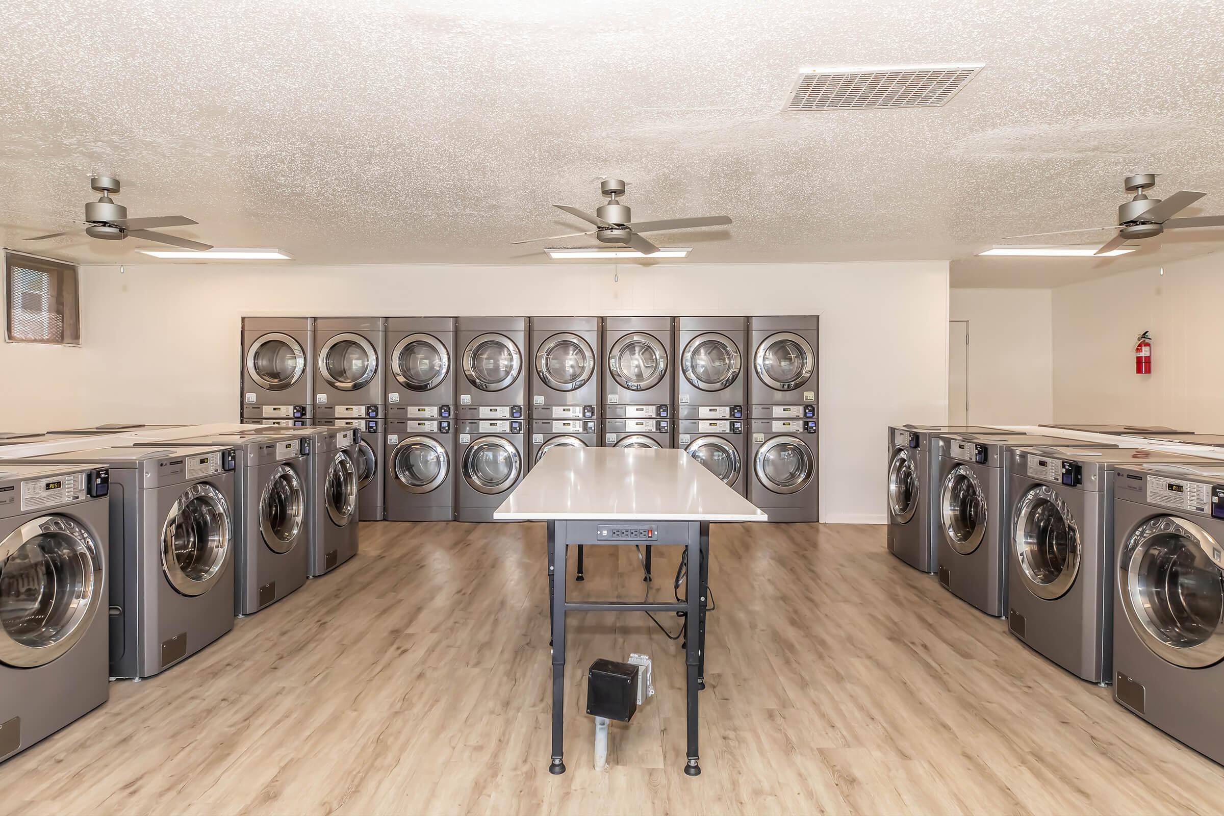 Modern laundry room featuring multiple industrial washing machines lined against the wall. The space is well-lit with ceiling fans and has a large folding table in the center, surrounded by a warm wood-like floor. The walls are painted a neutral color, creating a clean and organized environment.