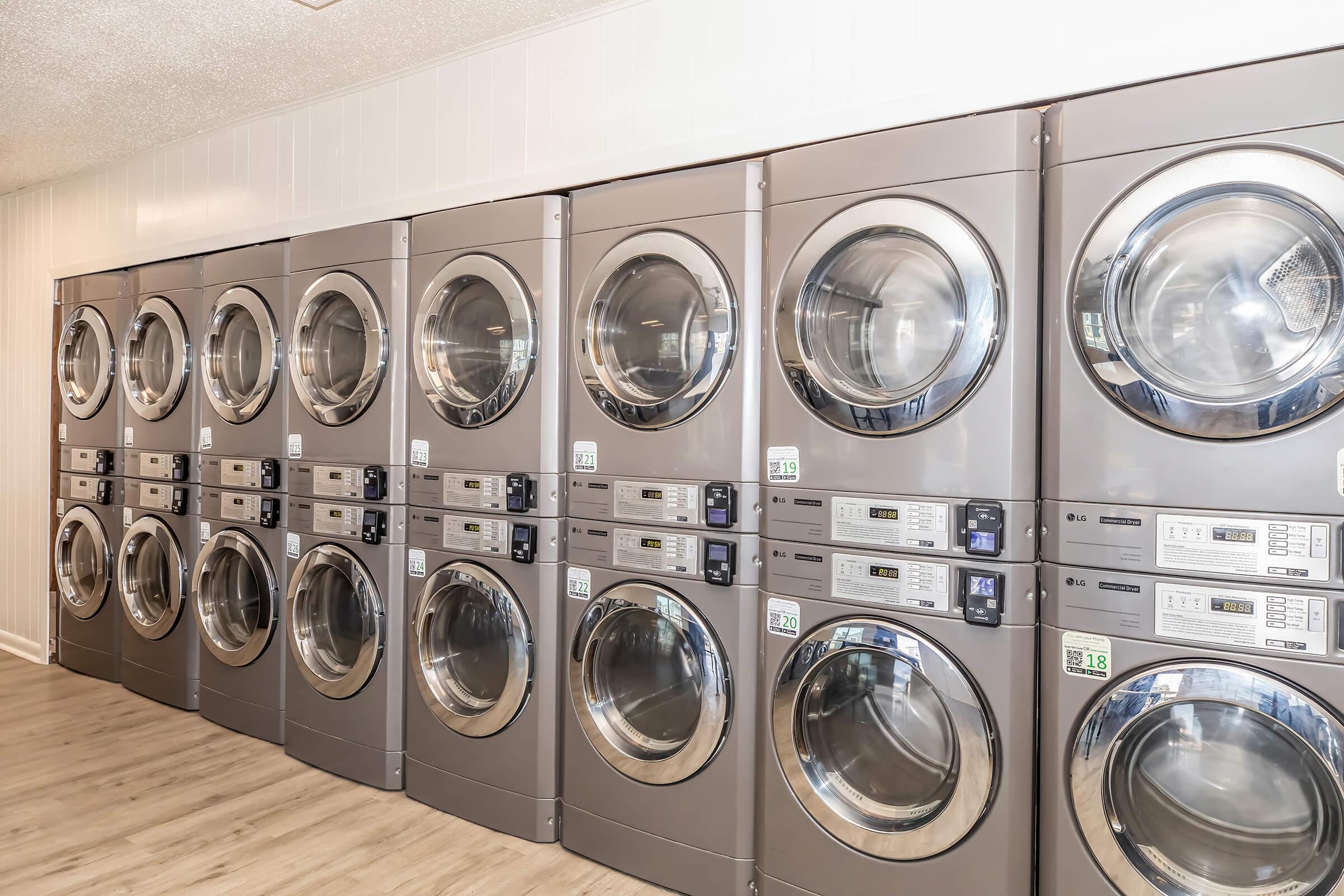 A row of modern, front-loading washing machines and dryers in a laundromat. The machines are silver with large glass doors, and they have digital control panels. The floor is wooden, and the walls are light-colored, creating a clean and organized laundry facility atmosphere.