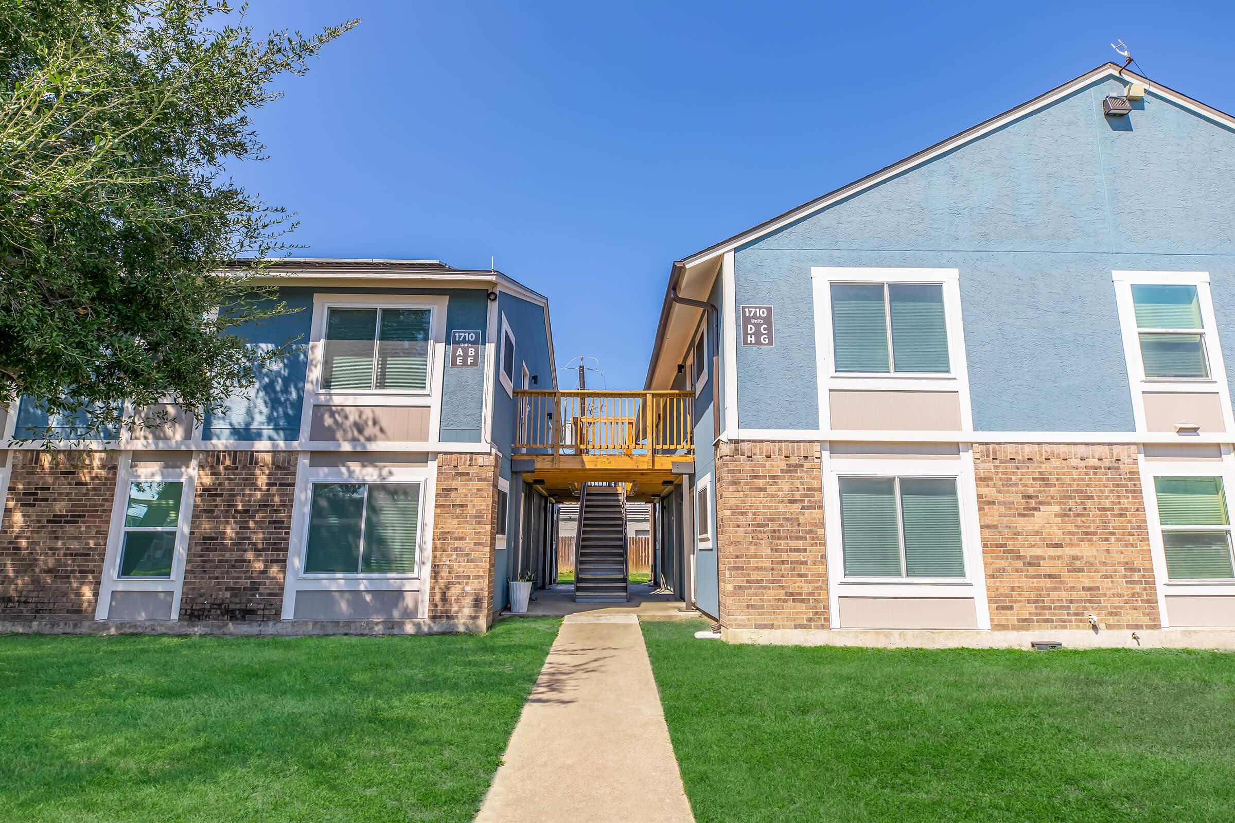 A view of two apartment buildings side by side, featuring a paved pathway leading to a raised deck with stairs. The buildings have a mix of blue and brick exteriors, and the surrounding area is grassy and well-maintained under a clear blue sky.