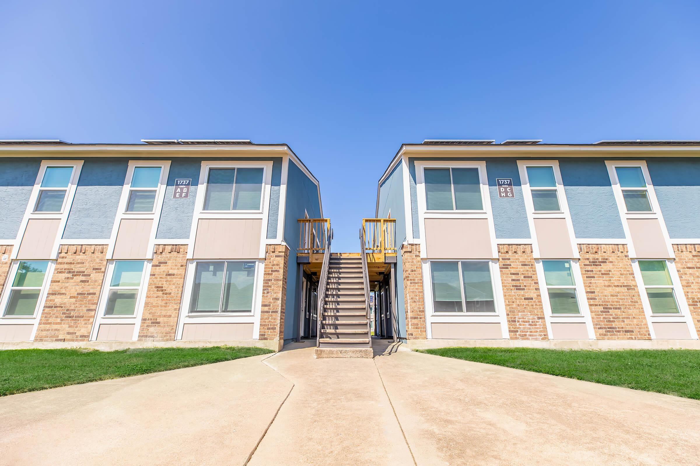 A two-story apartment building with a light blue and brick exterior. There is a central pathway leading to a set of stairs that connect the two sides of the building. Clear blue skies are visible above, and the grassy area in front of the building is neatly trimmed.