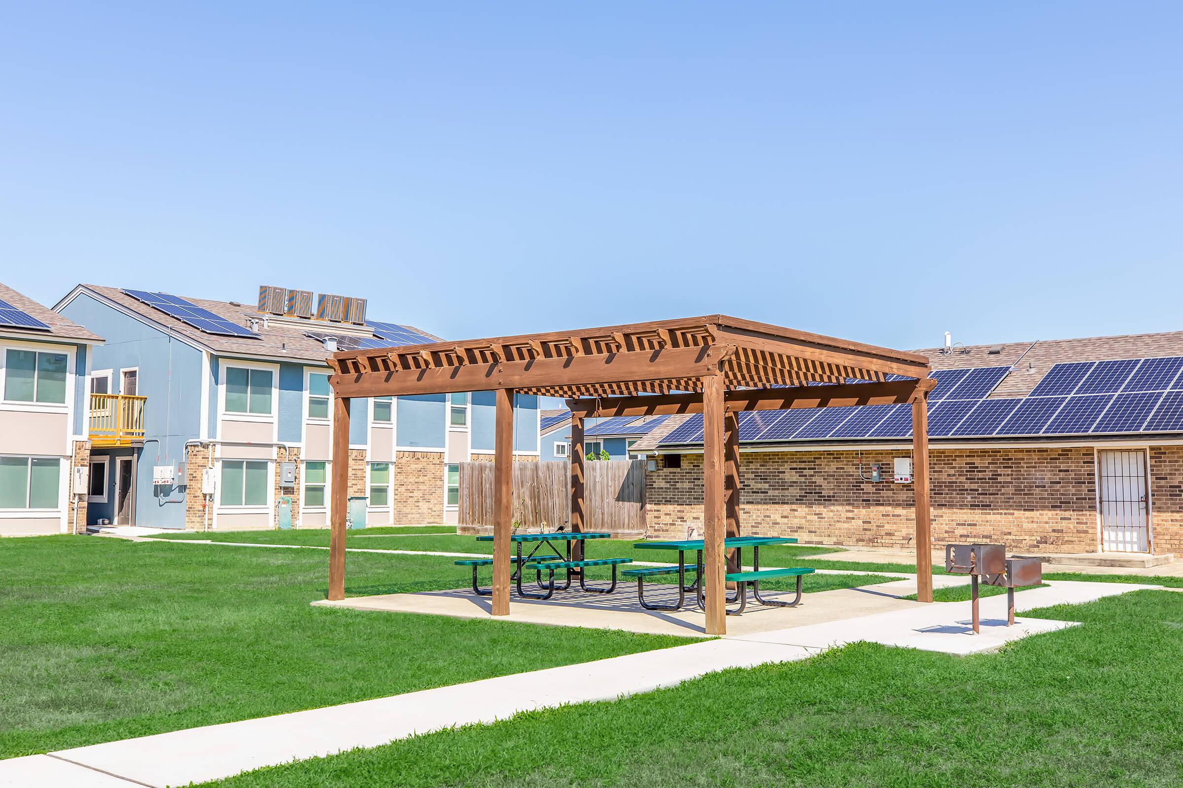 A wooden pergola with picnic tables underneath sits in a grassy courtyard surrounded by multi-story residential buildings. The buildings feature solar panels on their roofs, and the area is clear and sunny, creating a pleasant outdoor space for residents.