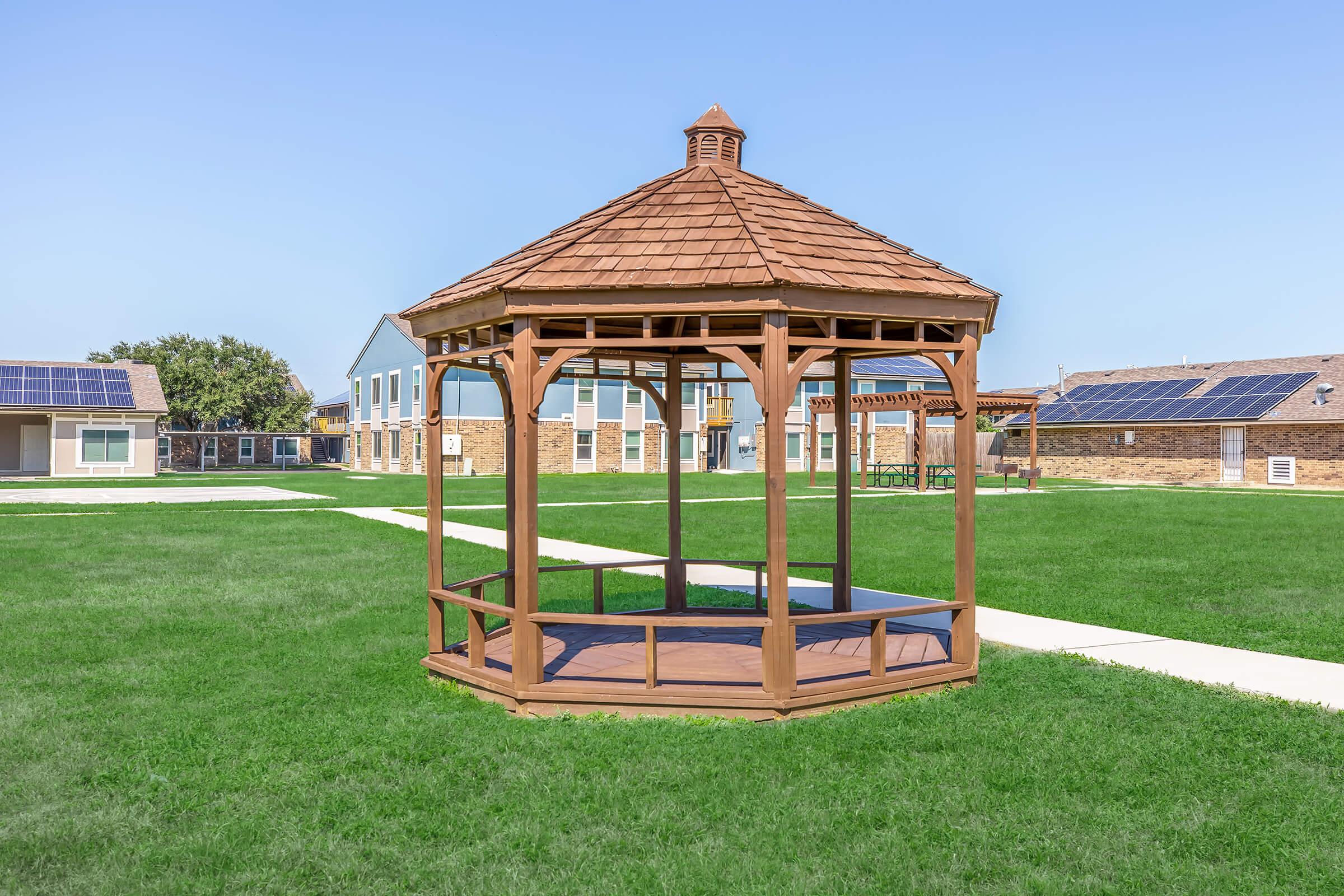 A wooden gazebo with a brown shingled roof, situated on a grassy lawn. In the background, there are residential buildings with solar panels on the roofs. The clear blue sky enhances the bright and open atmosphere of the outdoor space.