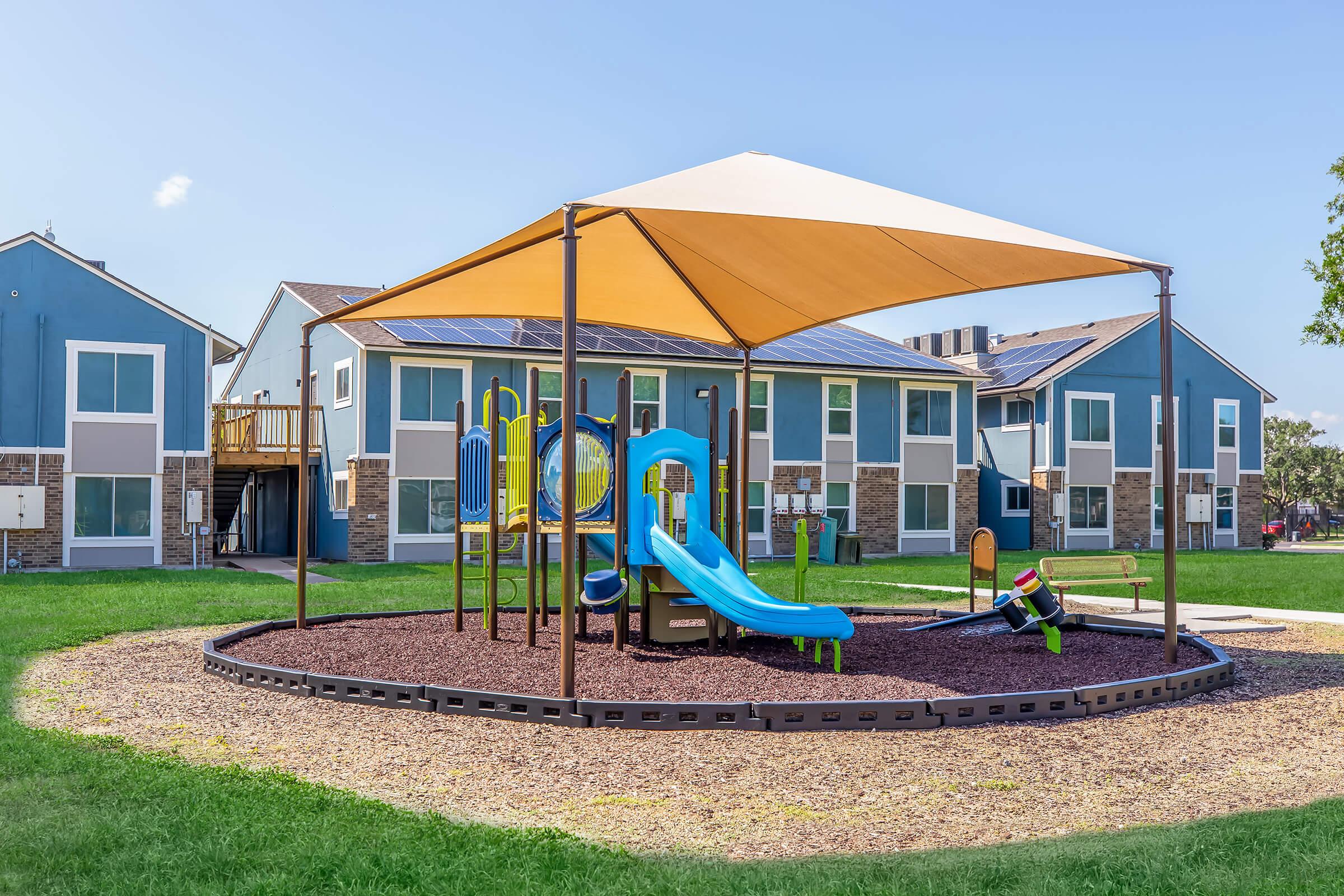 A colorful playground with a blue slide and various play structures, covered by a large shade canopy. Surrounding the playground is a mulch area, and in the background, there are blue and gray multi-unit residential buildings under a clear sky.