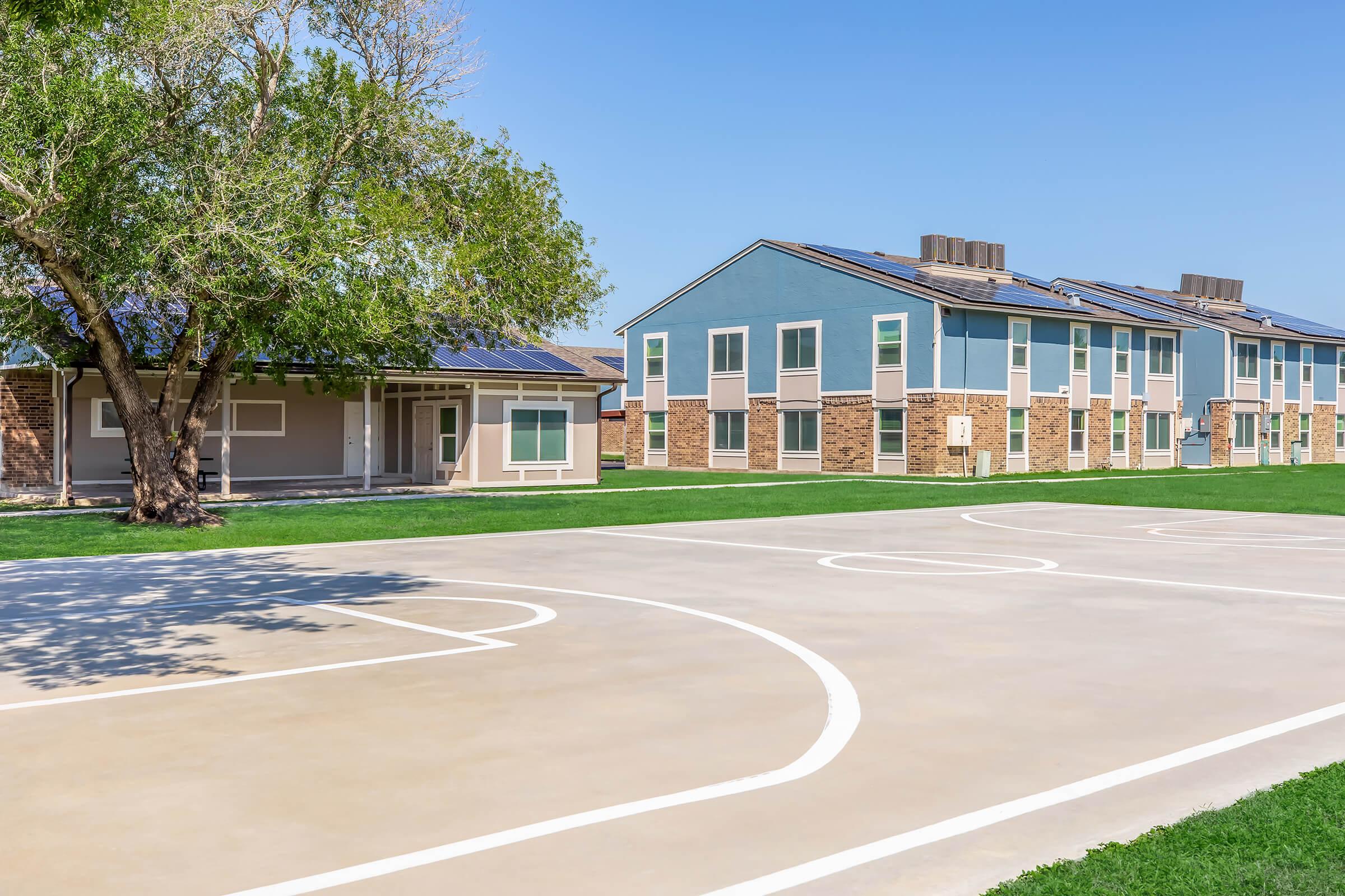 A basketball court with clean white lines in the foreground, surrounded by green grass. In the background, there are two modern residential buildings with solar panels on the roofs under a clear blue sky. A tree provides shade on one side of the court.