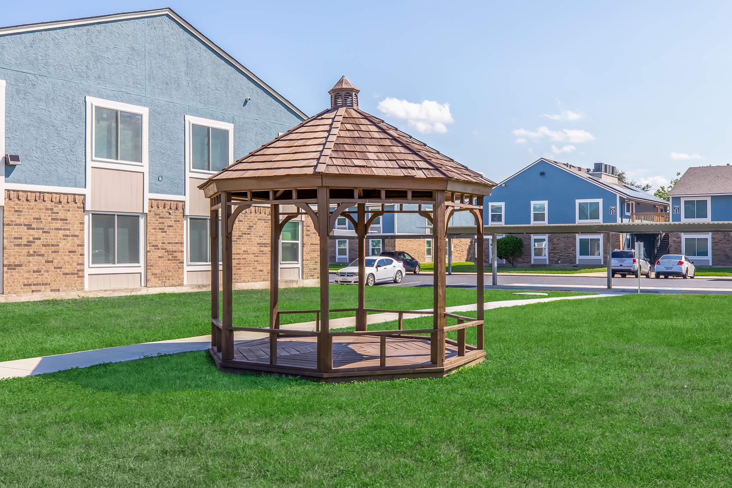 A wooden gazebo with a shingled roof stands on a green lawn, surrounded by a paved walkway. In the background, there are two multi-story buildings with a mix of brick and siding exteriors. A few parked cars are visible near the buildings under a clear blue sky.