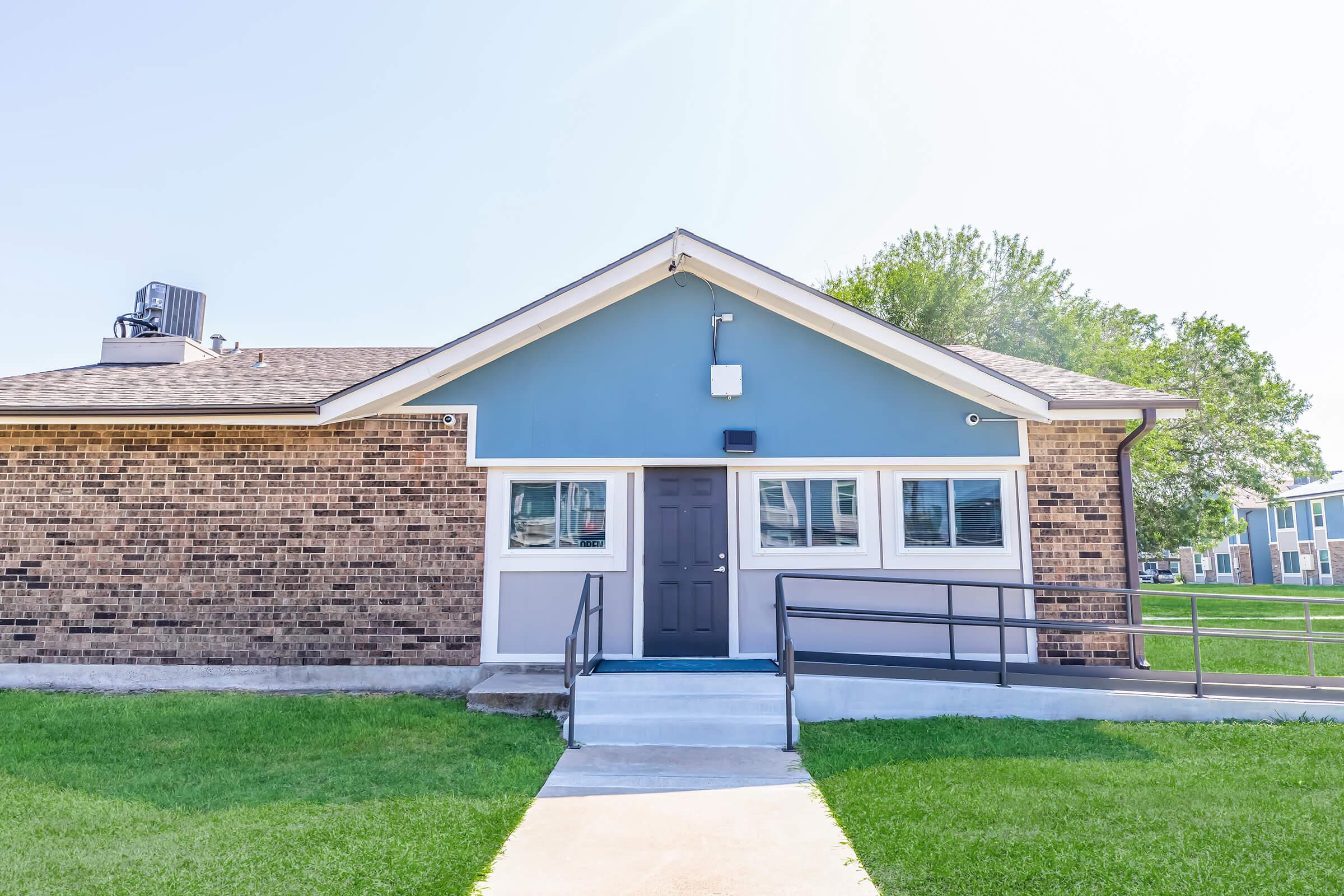 A single-story building with a blue and beige exterior, featuring a dark front door and multiple windows. A concrete walkway leads to the entrance, with a wheelchair-accessible ramp on the left. Lush green grass surrounds the building under a clear blue sky.