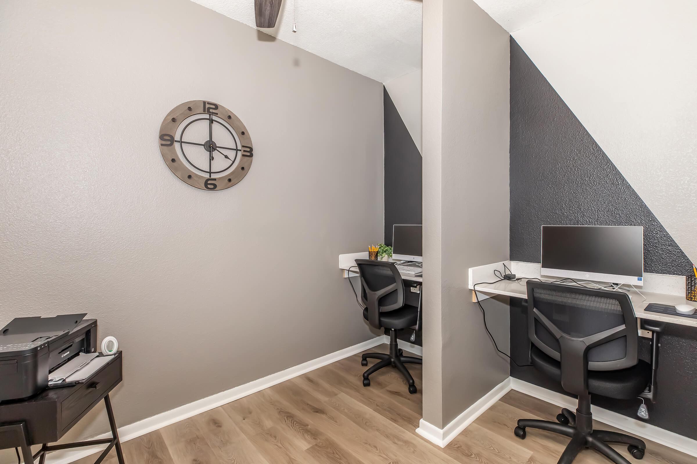 A small, modern home office space featuring two desks with computers, a printer on a side table, and a wall clock. The walls are painted in gray tones with one black accent wall, and there is a small plant on one of the desks. The flooring is light-colored wood.