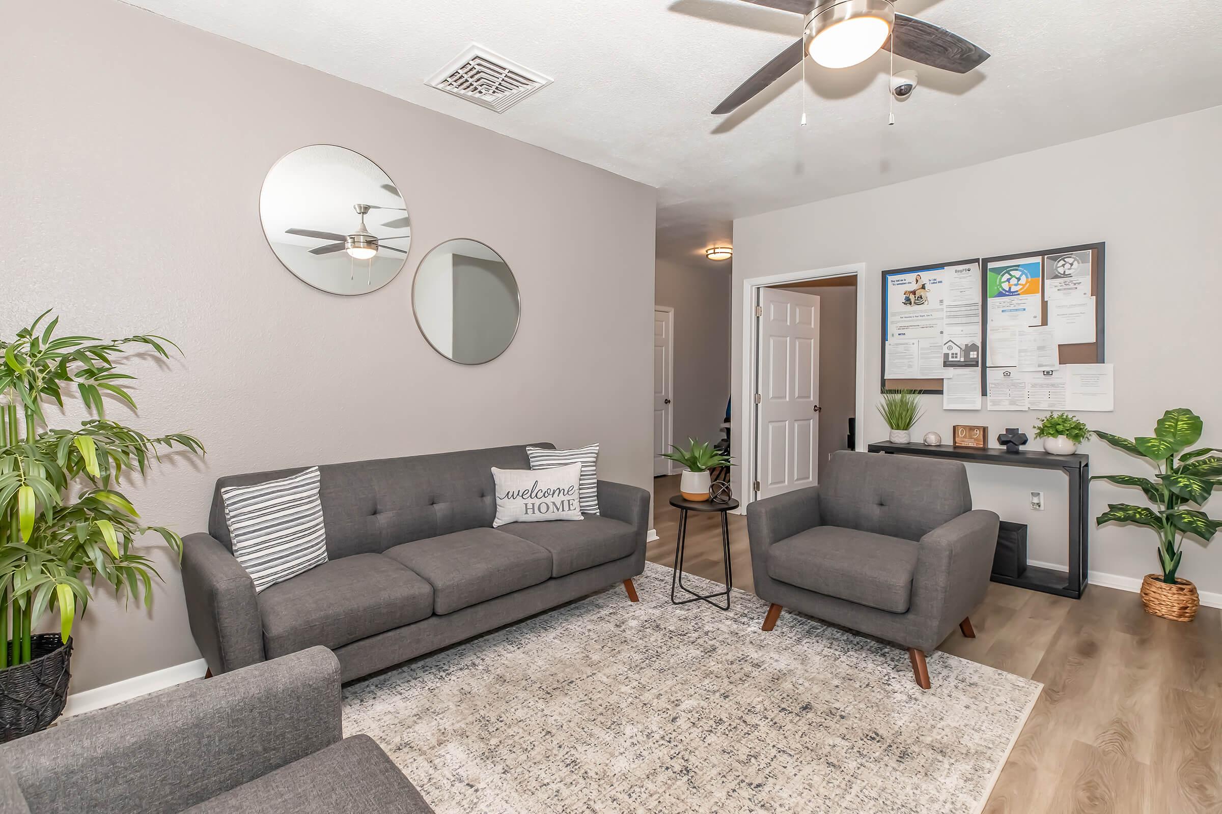A cozy living room featuring a grey sofa with a "welcome home" pillow, two matching armchairs, and a patterned area rug. There are two round mirrors on the wall, a ceiling fan, and potted plants adding greenery. A small table holds decorative items, and a bulletin board is visible in the background.