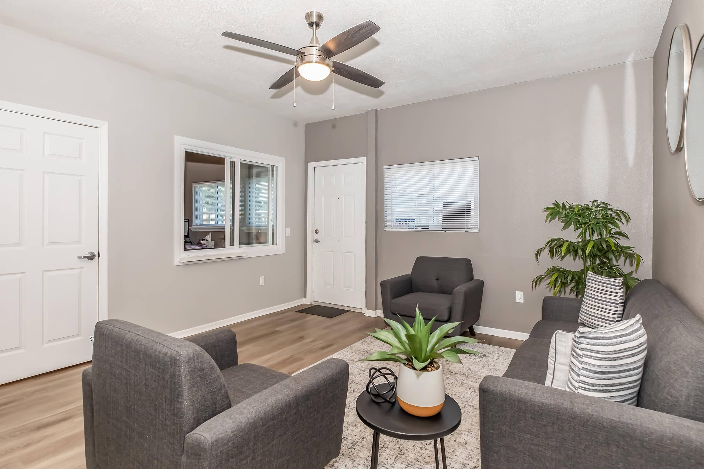 Modern living room featuring a gray sofa and two matching chairs, a small round black table, and a potted plant. A ceiling fan is above, and a front door and window provide natural light. The walls are painted a neutral color, and there is a decorative area rug on the floor.