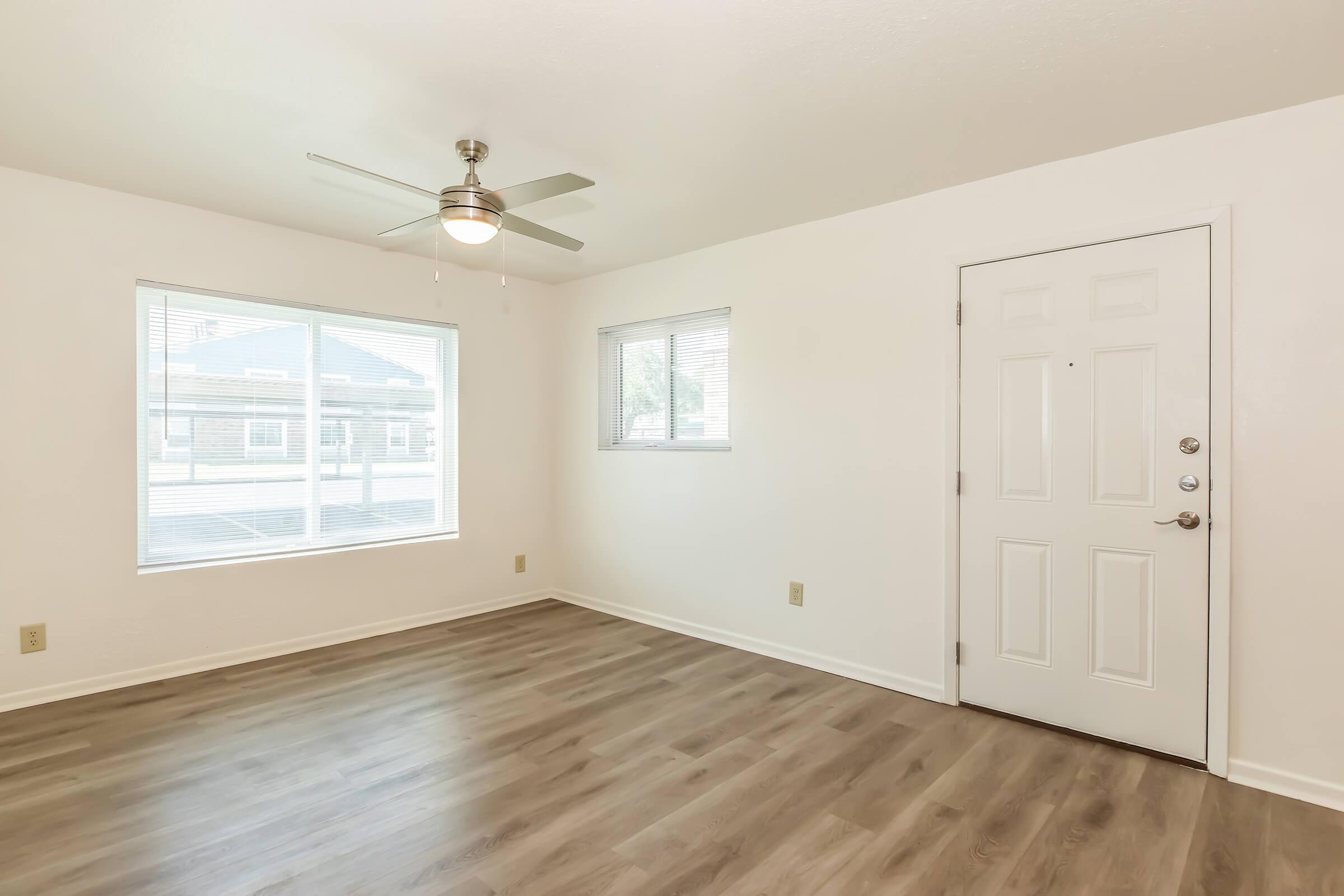 A bright, empty room featuring light-colored walls and wood-like laminate flooring. A ceiling fan is mounted on the ceiling, and there are two windows allowing natural light to fill the space. The room has a door on one wall, leading outside.