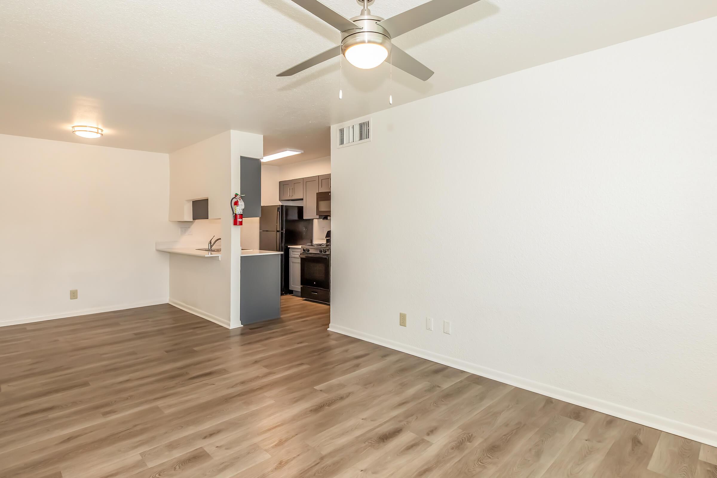 A spacious living area featuring light-colored walls and a ceiling fan. The open layout leads to a modern kitchen with dark cabinetry and appliances. The flooring is a warm, wood-like material, creating a cozy atmosphere. There's ample natural light and room for furniture arrangement.
