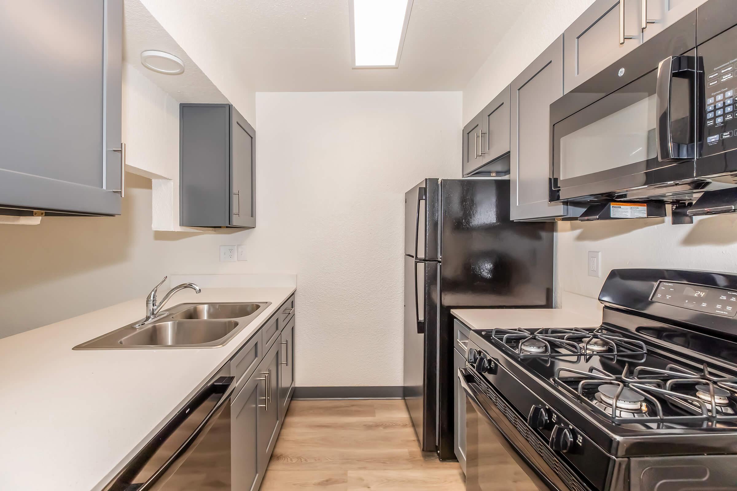 Modern kitchen featuring stainless steel appliances, including a gas stove, microwave, double sink, and a black refrigerator. The cabinetry is dark gray, and the countertops are light-colored. The space is well-lit with overhead lighting and has a clean, contemporary design.
